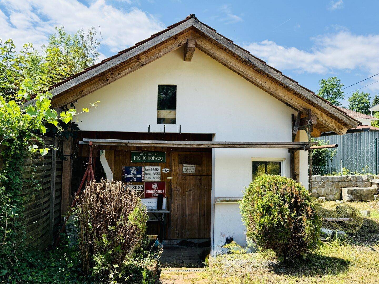 Kleines Nebengebäude im Garten mit Holztür und Satteldach, umgeben von Pflanzen.