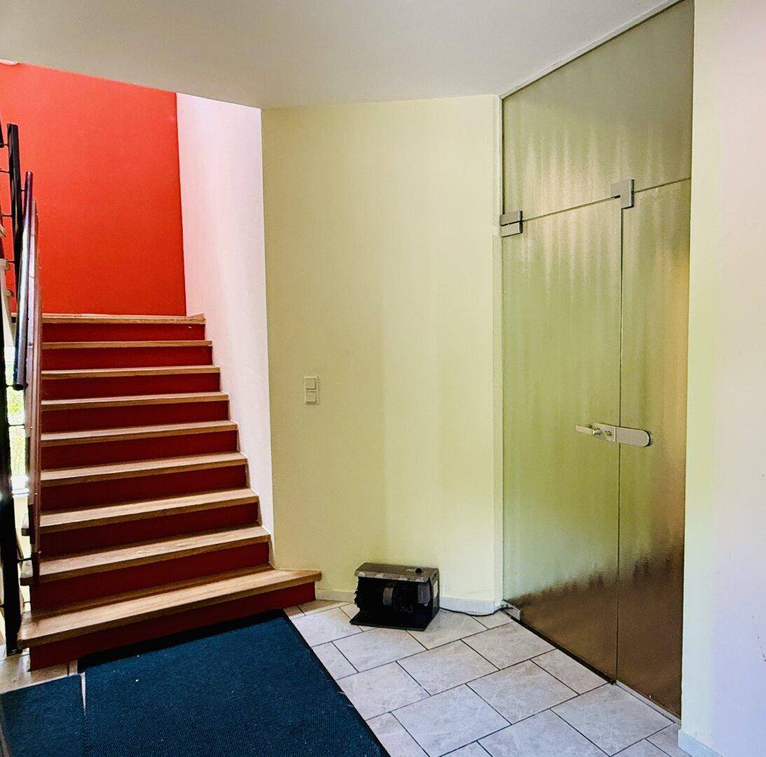 Heller Eingangsbereich mit gefliestem Boden, einer Holztreppe und einer roten Akzentwand.