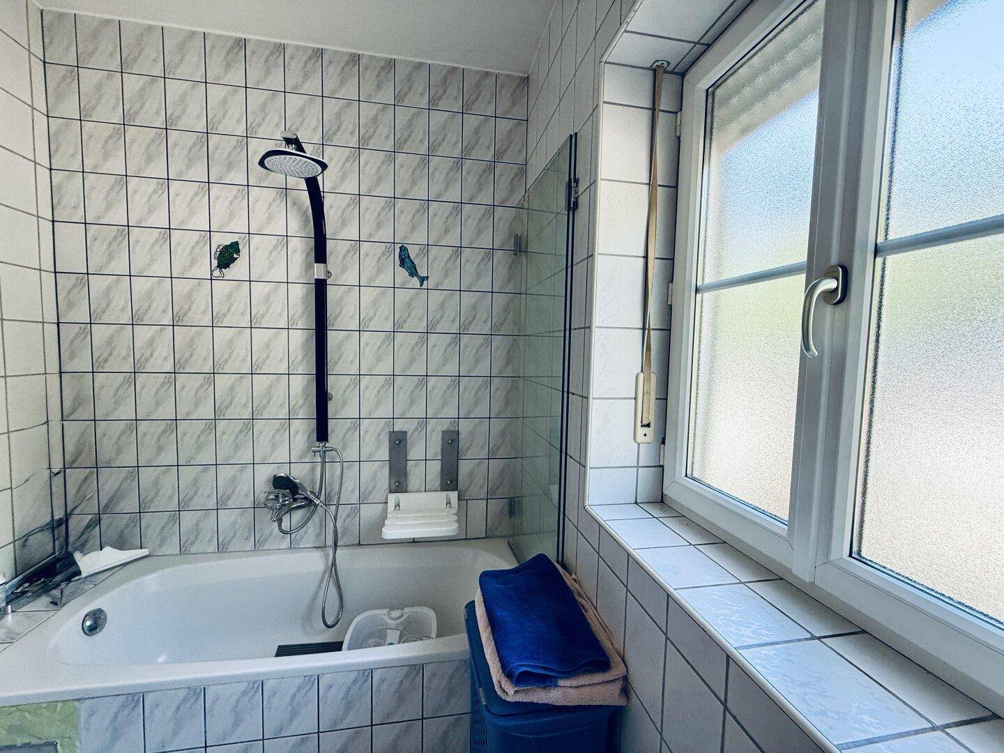 Badezimmer mit Badewanne, Duschvorrichtung und weißen Fliesen, hell durch ein Fenster.