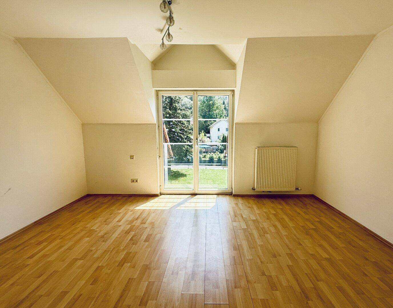 Geräumiges Dachzimmer mit Holzfußboden und großer Fenstertür mit Blick ins Grüne.
