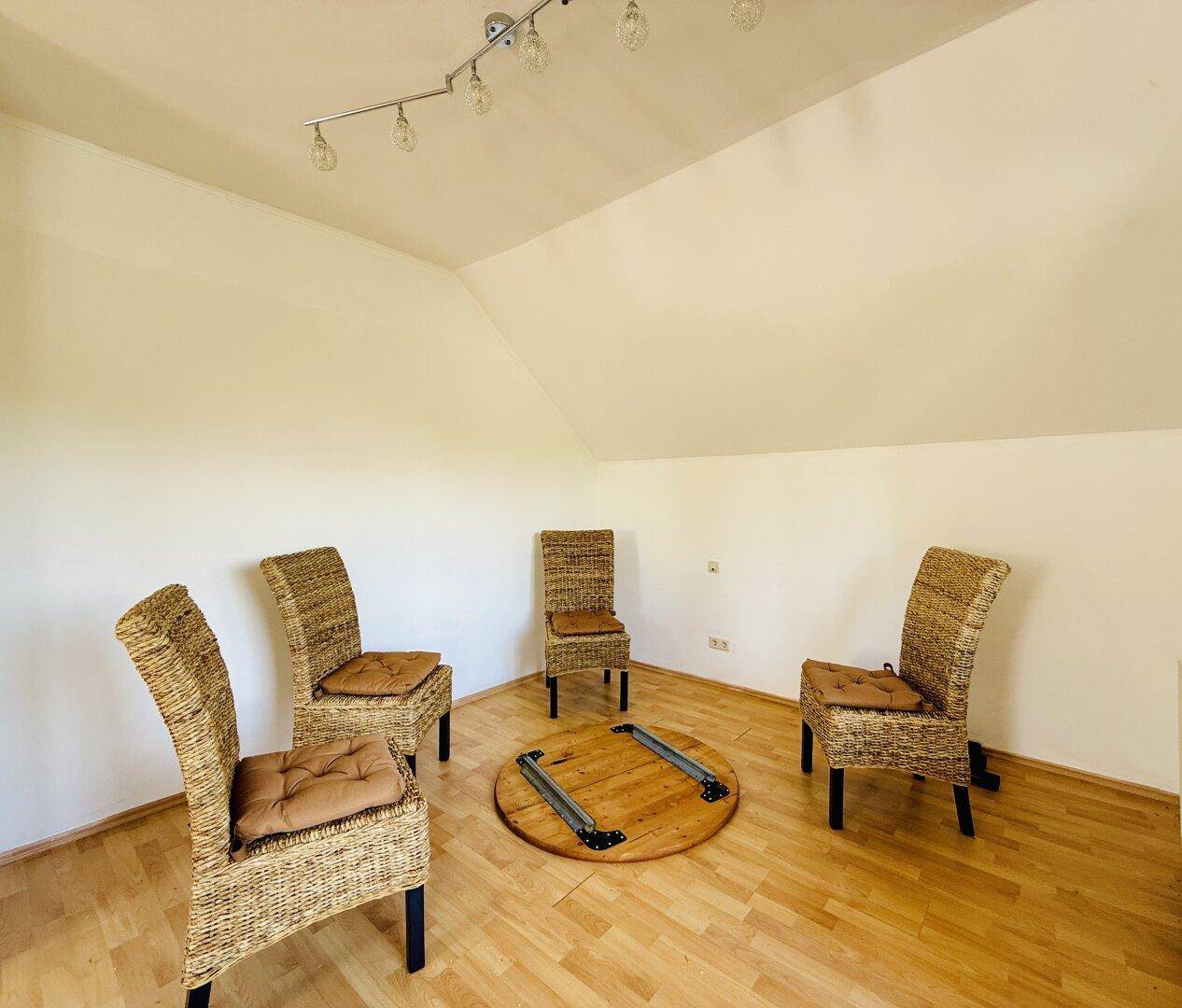 Dachgeschosszimmer mit Holzfußboden, Korbstühlen und einem runden Holztisch.