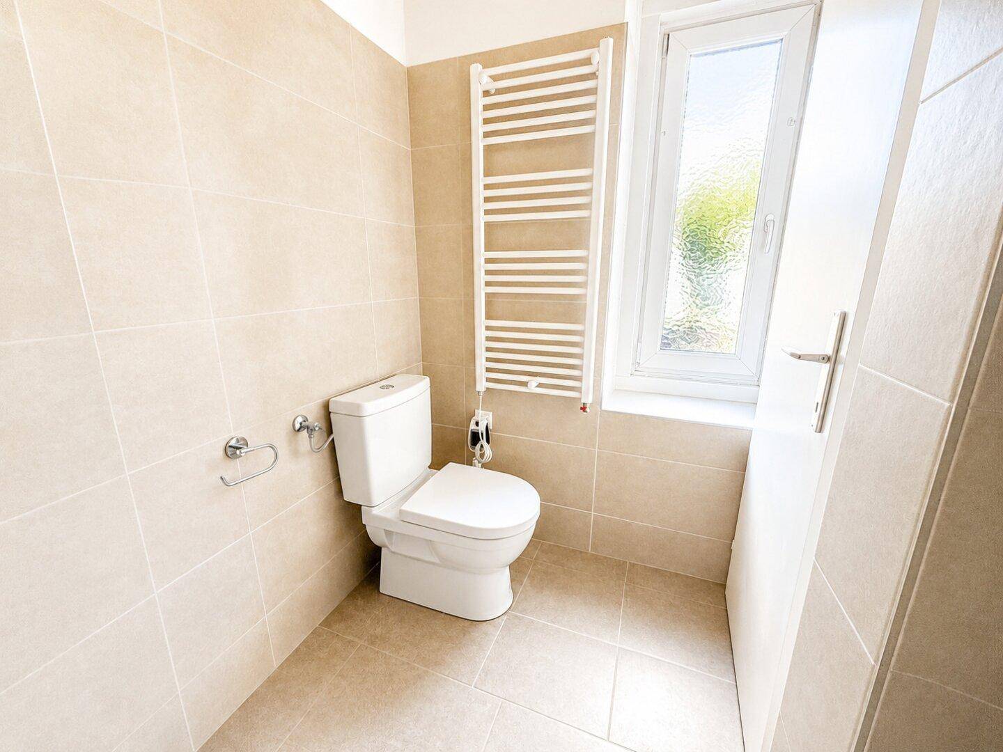 Helles Badezimmer mit WC, Fliesenboden und beheiztem Handtuchhalter am Fenster.