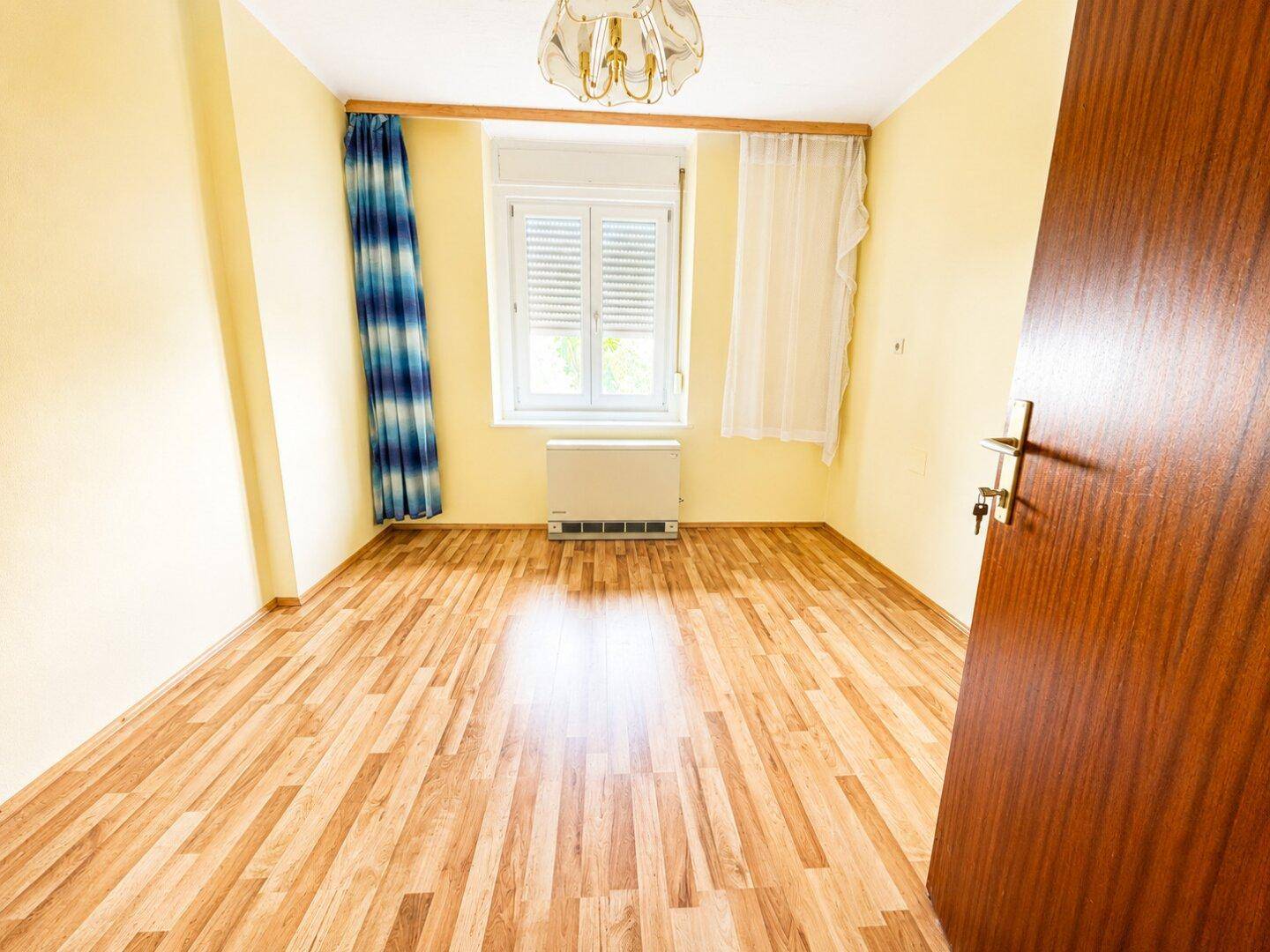 Leeres Zimmer mit Laminatboden, Fenster mit Jalousien und gelben Wänden.