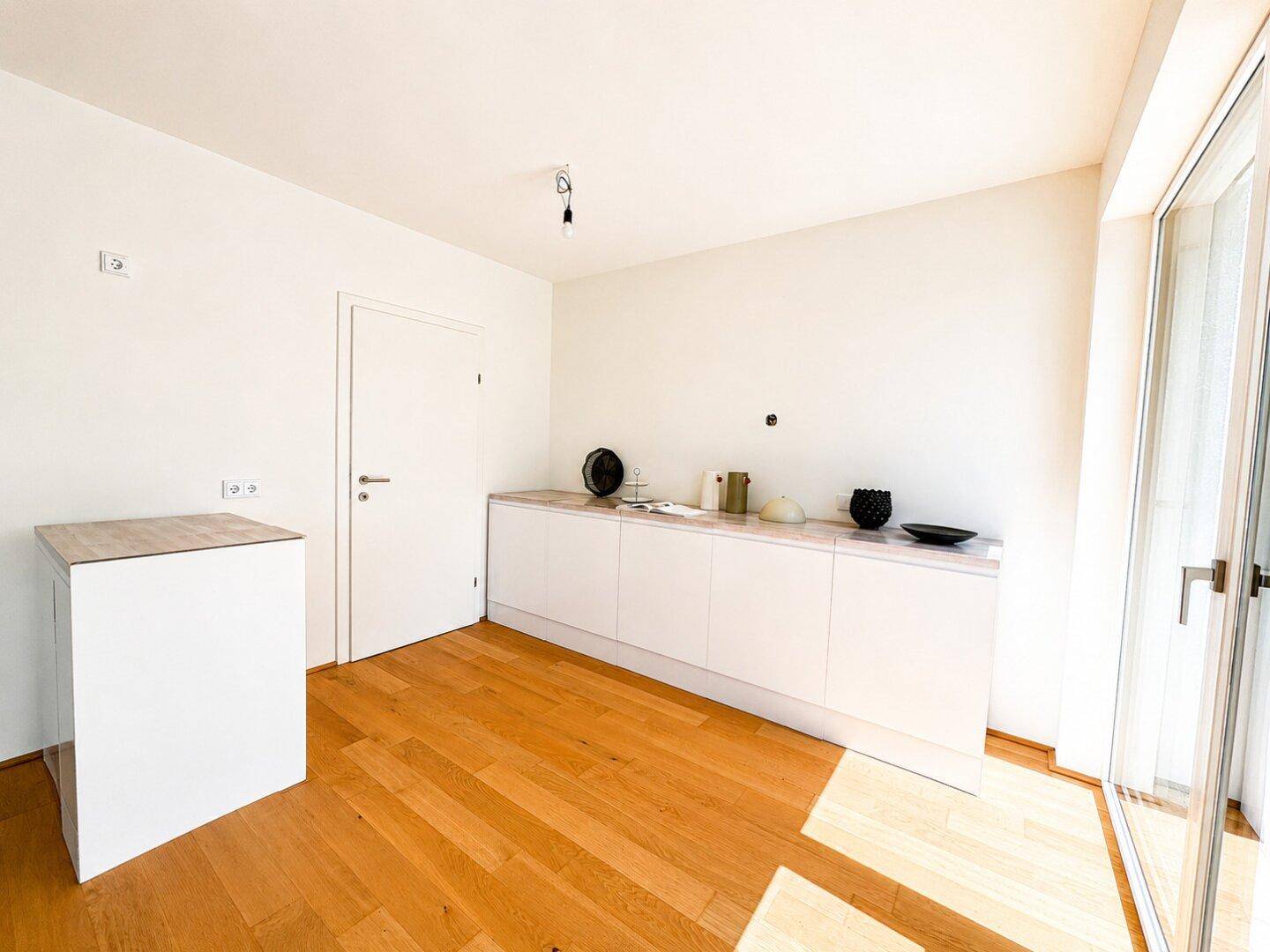 Moderne Küche mit weißen Schränken, Holzarbeitsplatte und viel Tageslicht durch große Fenster.
