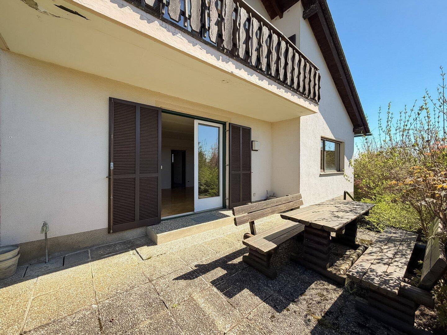 Sonnige Terrasse mit rustikalen Holzmöbeln und Zugang zum Haus, darüber ein Balkon.