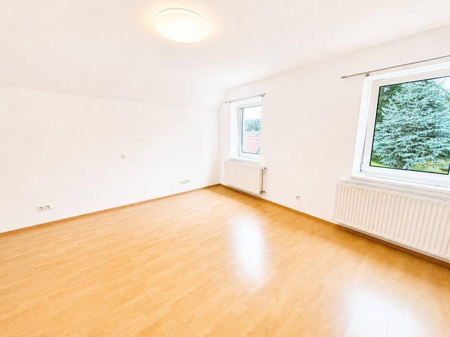 Geräumiges Schlafzimmer mit hellem Holzboden und Blick ins Grüne durch die Fenster.