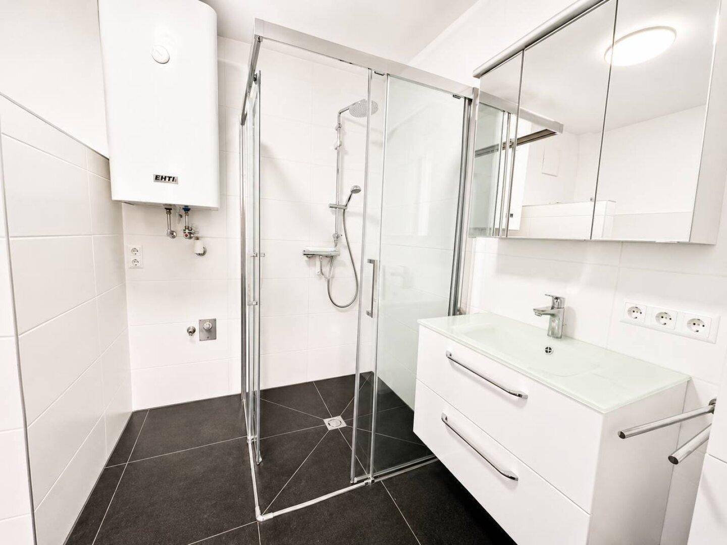Modernes Badezimmer mit Duschkabine, weißem Waschtisch und dunklen Bodenfliesen.