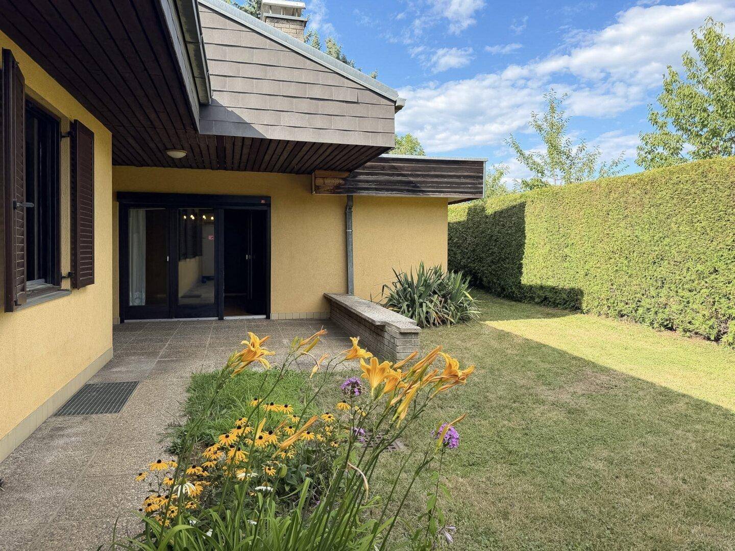 Terrasse mit Zugang zum Garten, umgeben von Blumen und einer hohen Hecke.