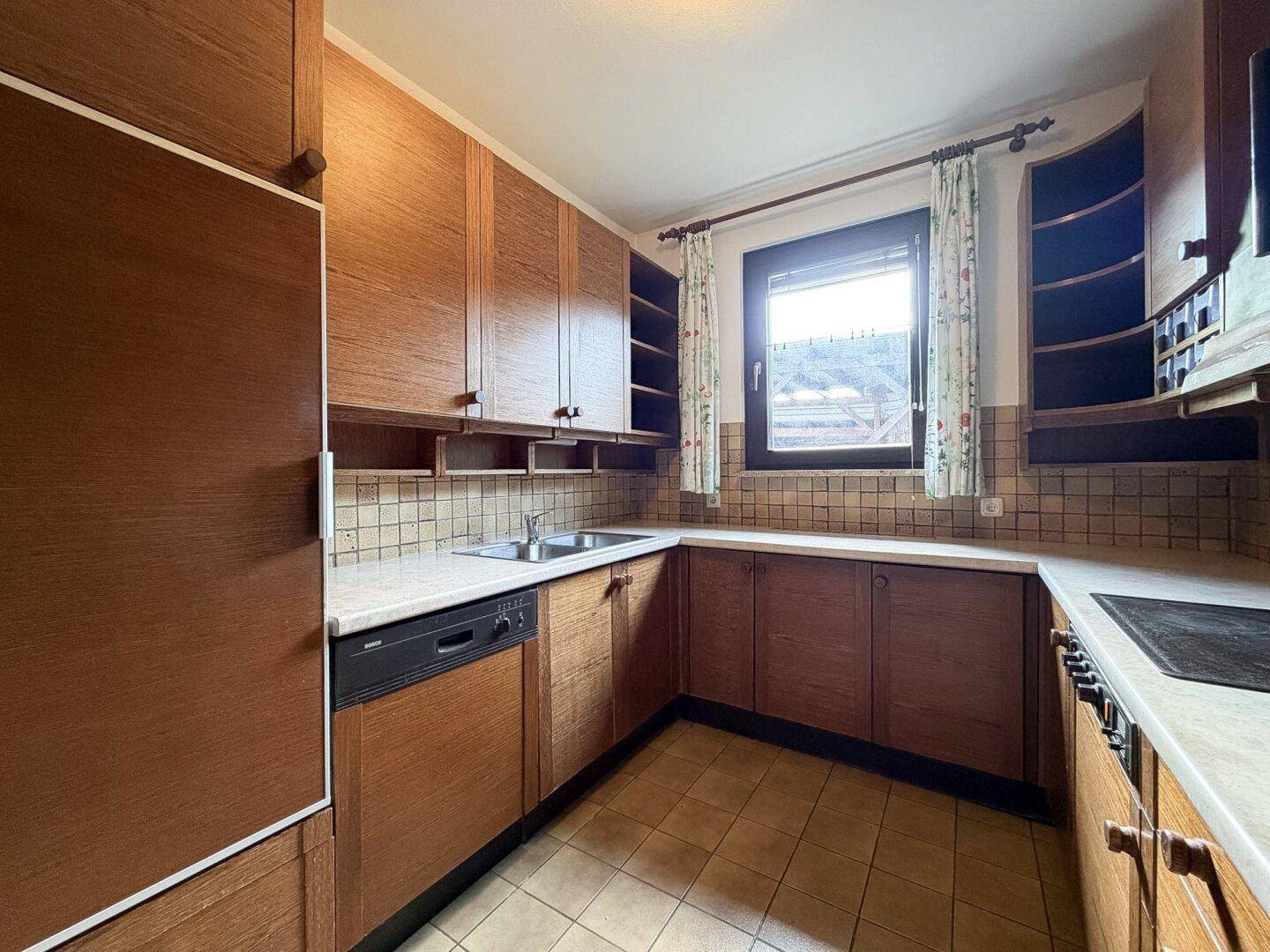 Funktionale Küche mit Holzschränken, Fliesenboden und Fenster mit Blick ins Grüne.