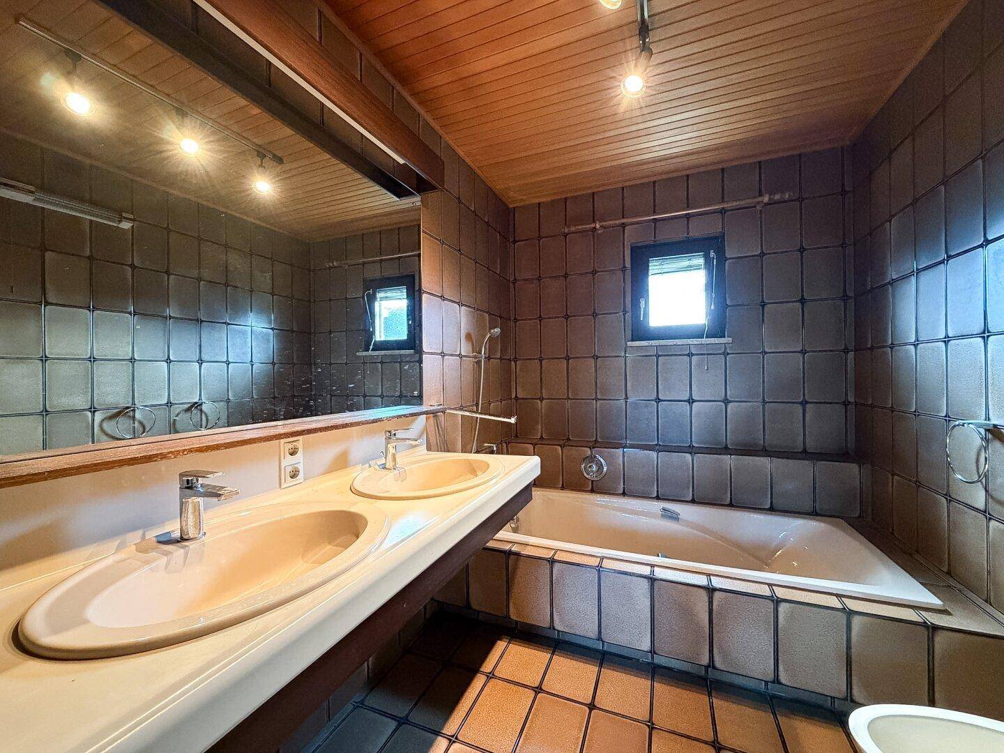 Badezimmer mit Doppelwaschbecken, Badewanne und braunen Fliesen im 80er-Jahre-Stil.