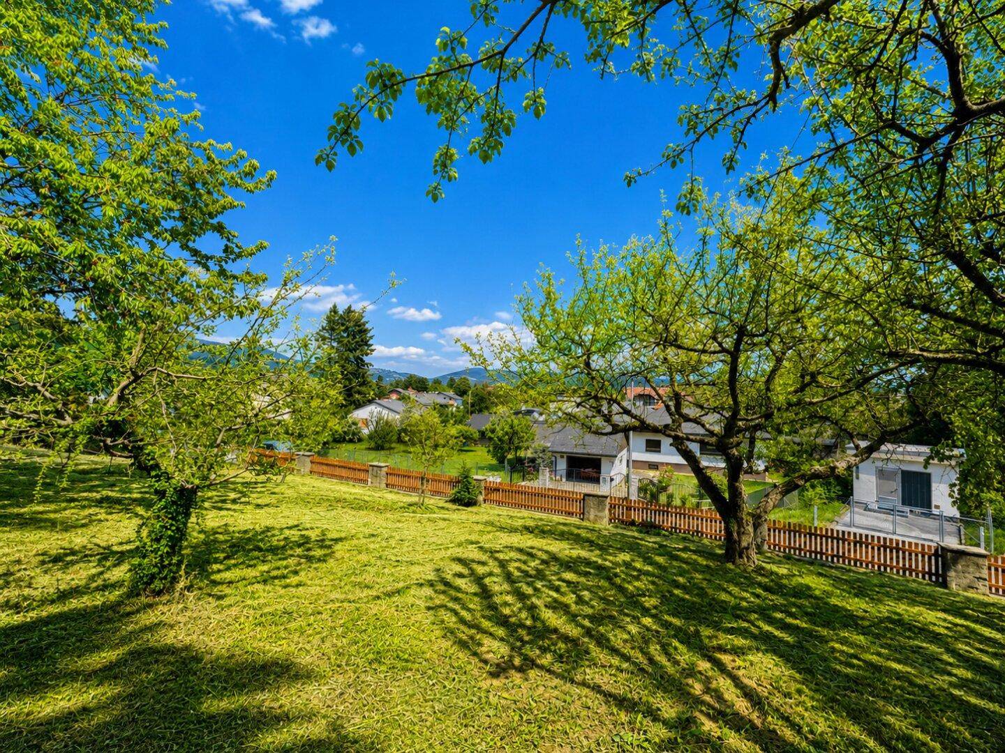Sonniger Garten mit Rasen, Bäumen und einem malerischen Blick auf die umliegende Natur und Häuser.