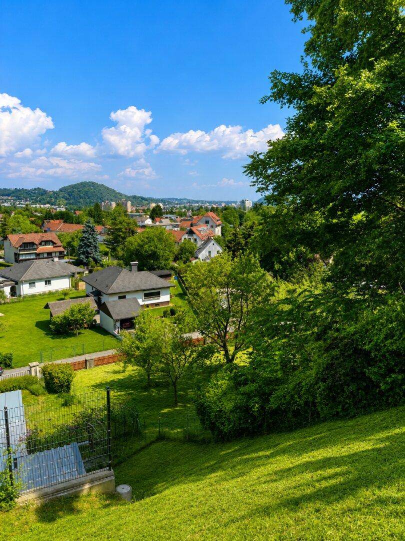 Panoramablick über eine grüne Hanglage mit Bäumen und einer Stadt im Hintergrund unter blauem Himmel.