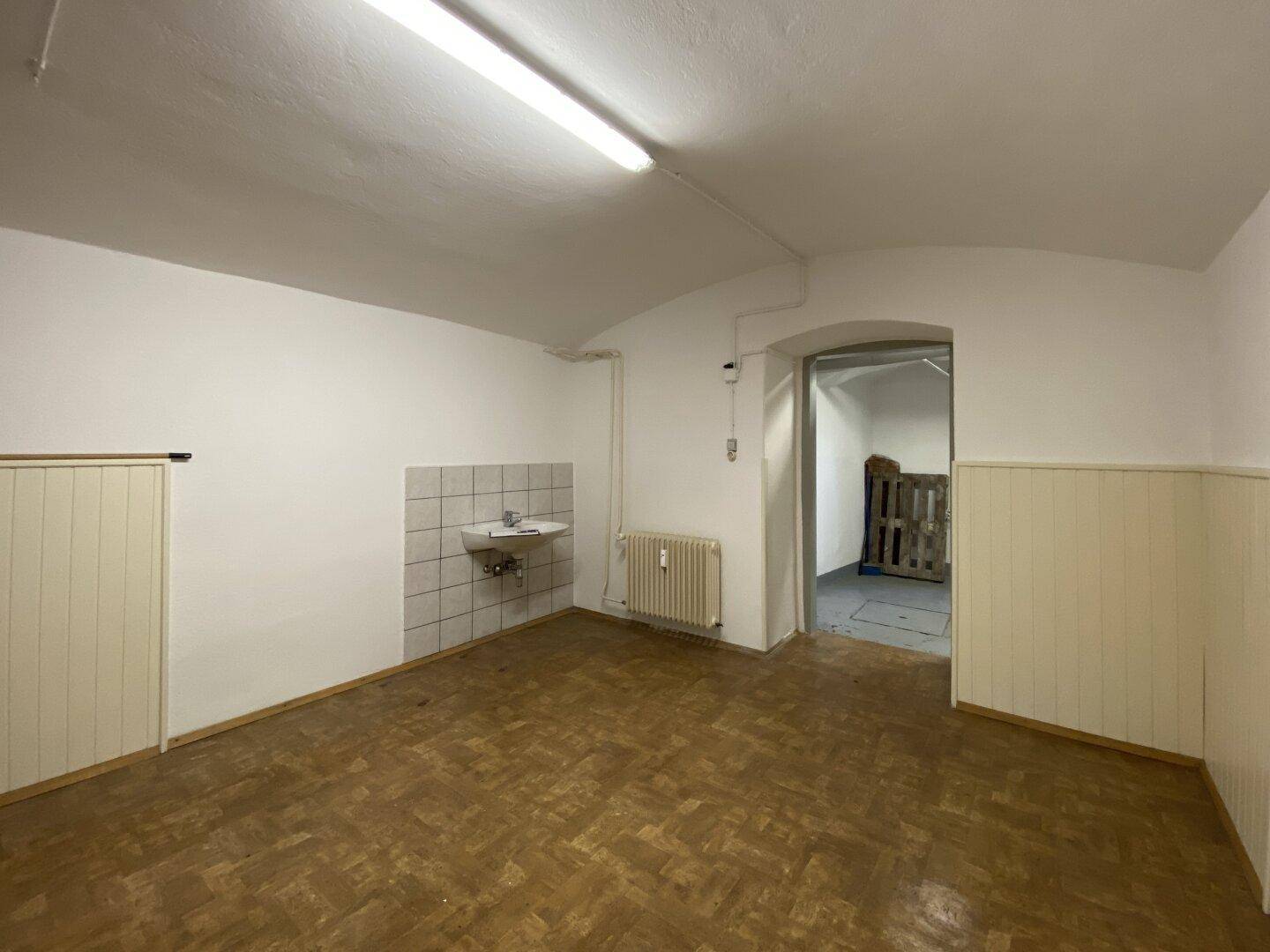 Kellerraum mit Waschbecken, Heizkörper und Blick in einen angrenzenden Lagerbereich.