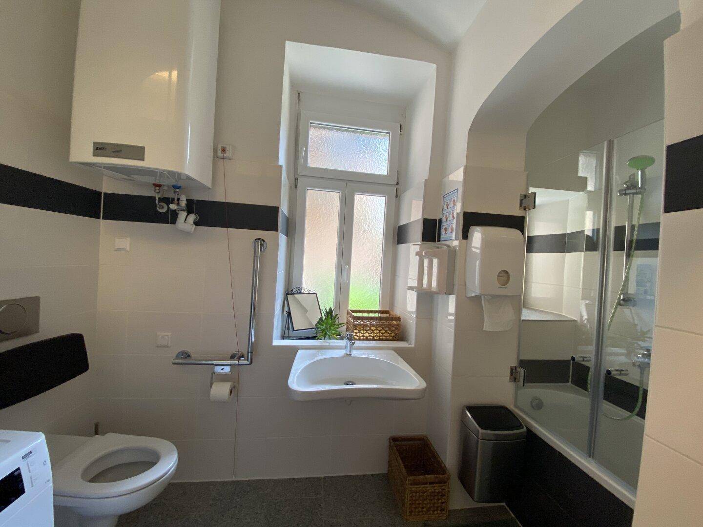 Badezimmer mit weißen und schwarzen Fliesen, Dusche, WC, Waschbecken und Fenster, ausgestattet mit Haltegriffen.