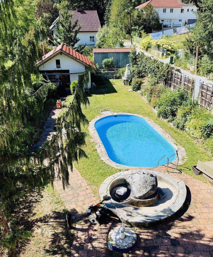 Blick von oben auf den Garten mit Swimmingpool, Nebengebäude und dekorativem Steinbrunnen.