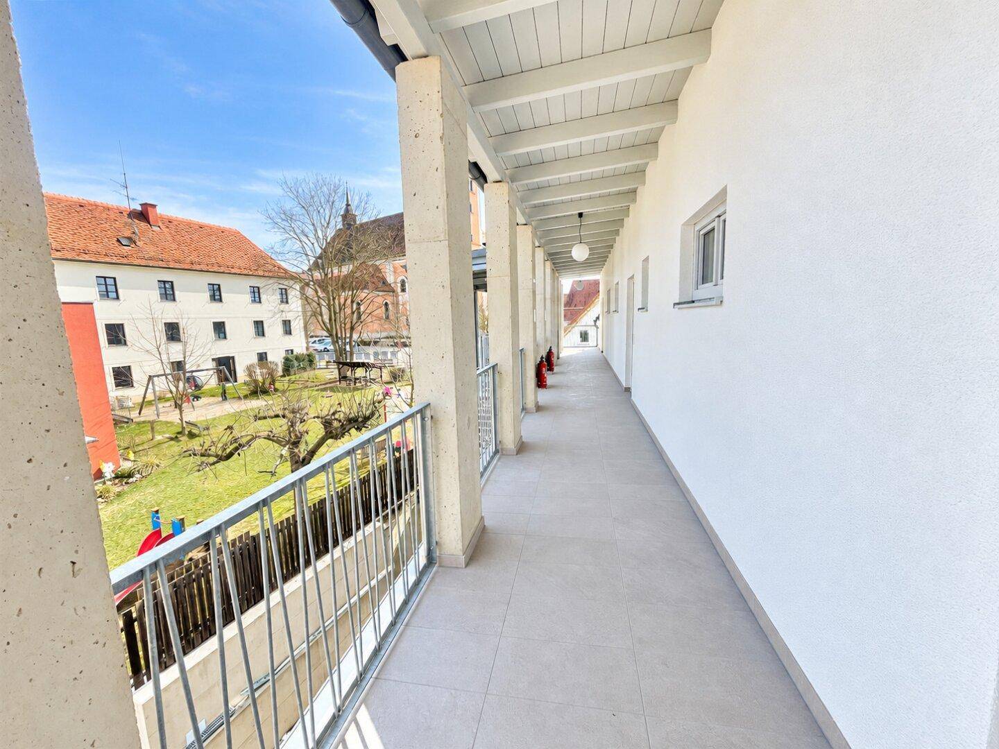 Lange, überdachte Loggia mit Blick auf einen grünen Innenhof und Spielplatz.