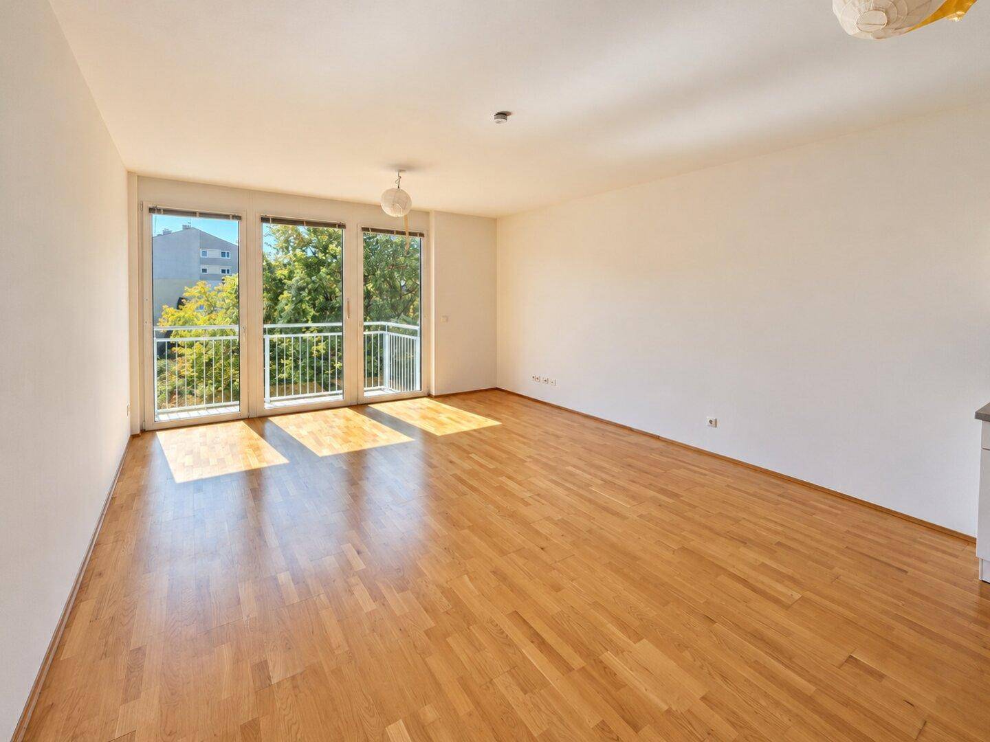 Helles, leeres Wohnzimmer mit großen Fenstern, Holzboden und Zugang zum Balkon.