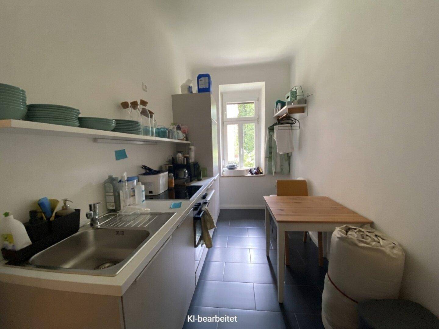 Schmale Küche mit grauen Schränken, dunklem Fliesenboden und einem kleinen Esstisch am Fenster.