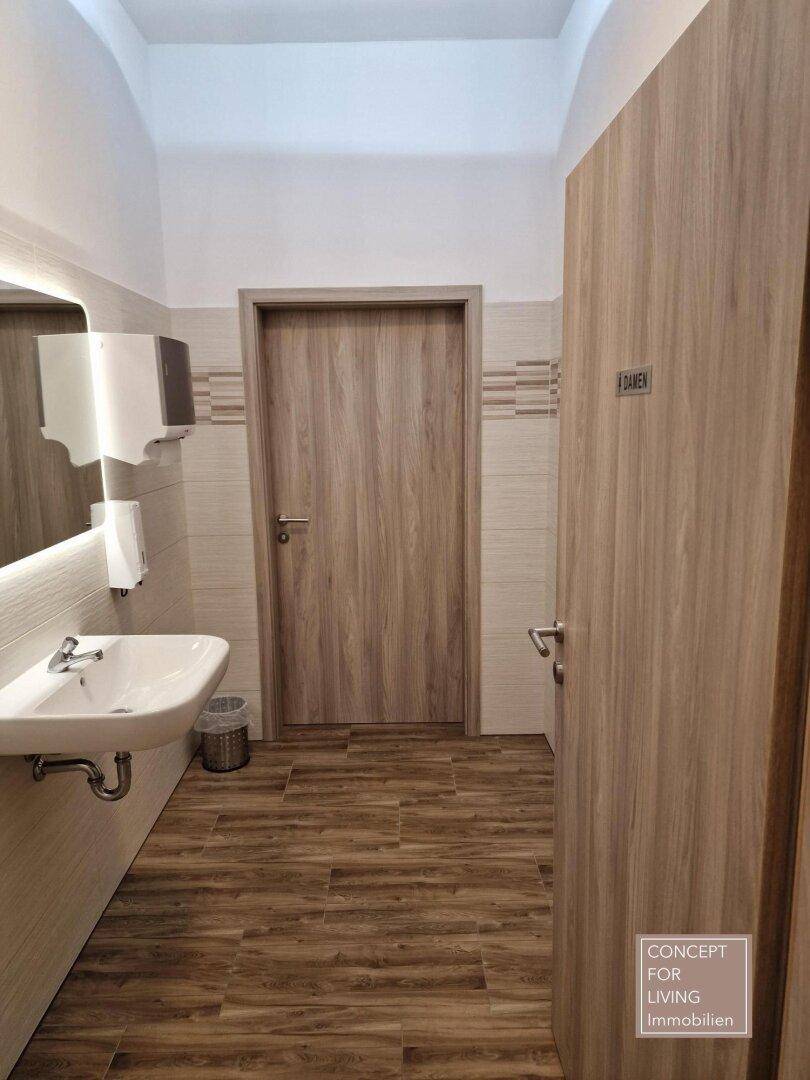 Modernes Damen-WC mit Waschbecken, großem Spiegel und Holzoptik-Fliesenboden.