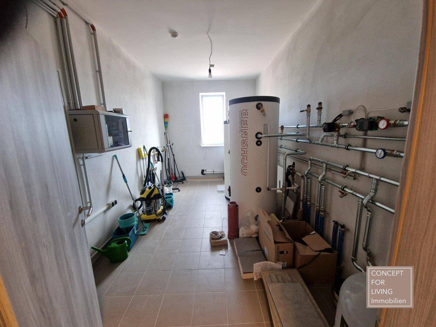 Technikraum mit Warmwasserspeicher, umfangreicher Rohrinstallation und einem kleinen Fenster.