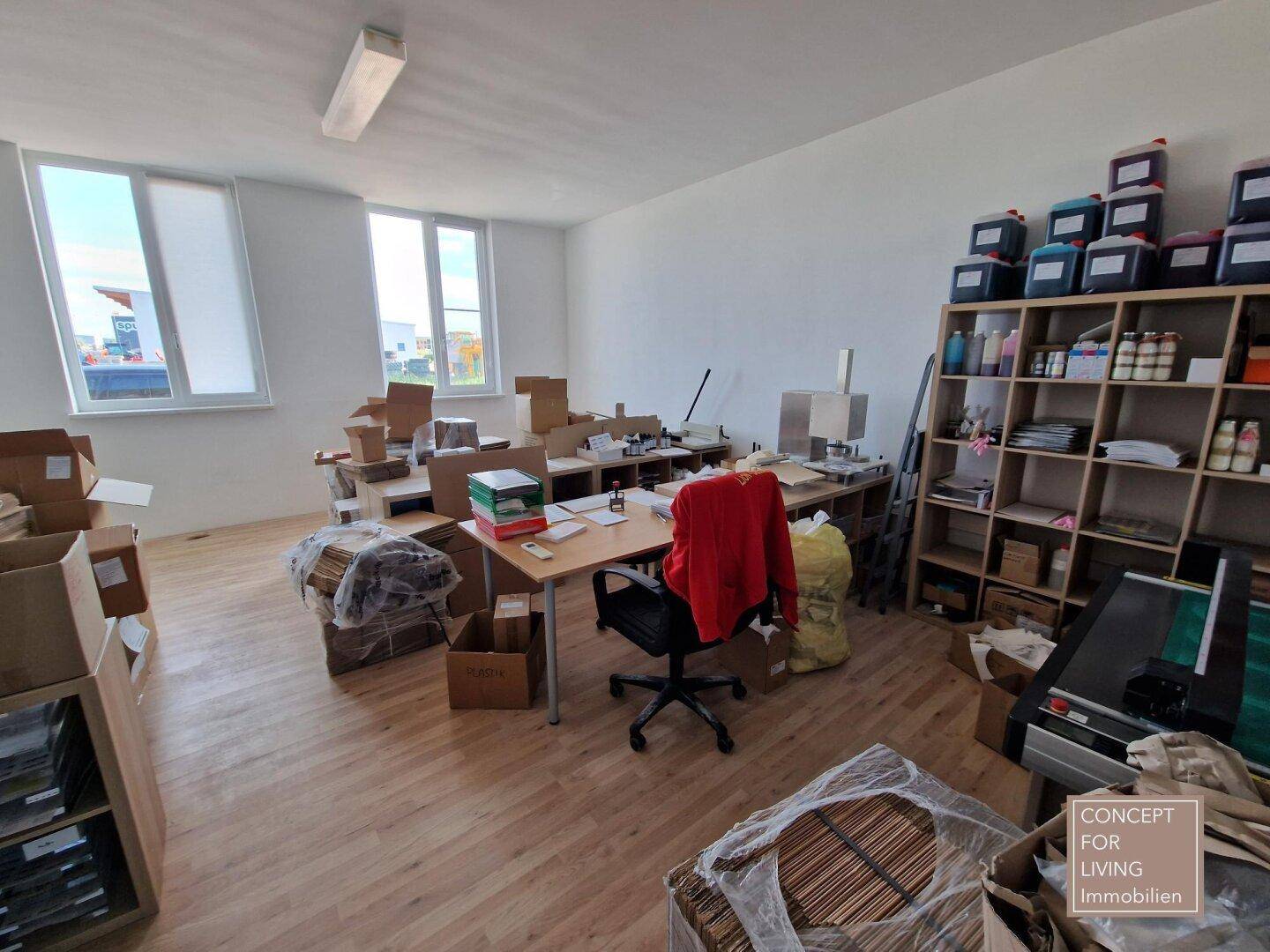 Großes Büro mit mehreren Arbeitsplätzen, Regalen und Kartons, gut beleuchtet durch Fenster.