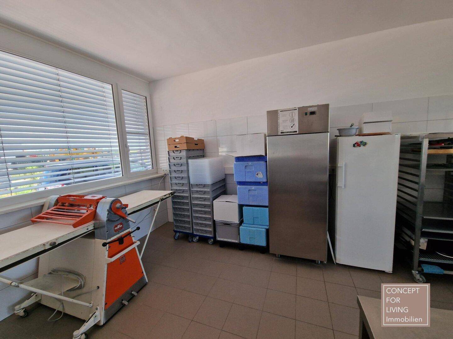 Lagerbereich in der Gewerbeküche mit Kühlgeräten, Regalen und einem großen Fenster mit Jalousien.