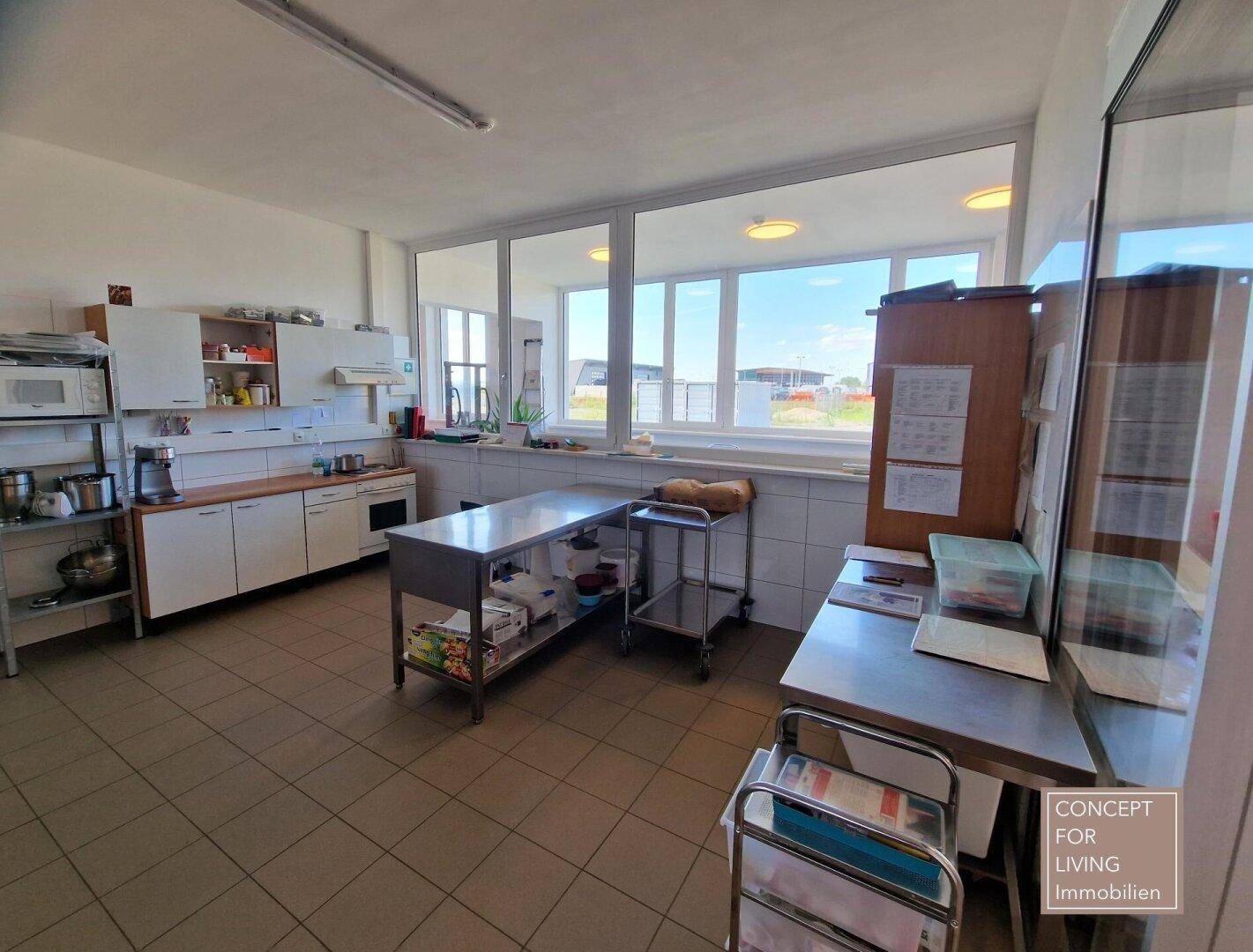Helle Gewerbeküche mit umfangreichen Arbeitsflächen, Regalen und Blick nach draußen durch große Fenster.