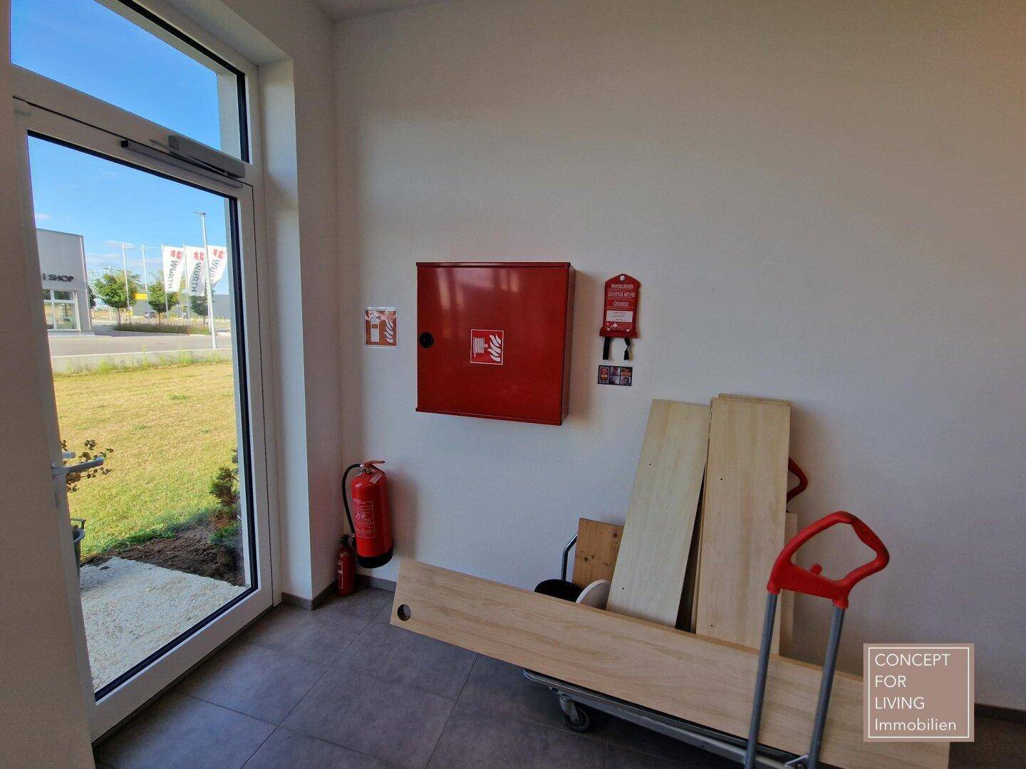 Eingangsbereich mit Feuerlöscher und Brandschutzschrank, neben einer großen Glastür mit Blick nach draußen.