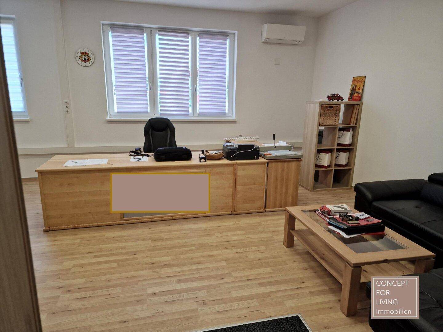 Helles Büro mit großem Schreibtisch, schwarzem Ledersofa und Holzboden, gut beleuchtet durch Fenster.