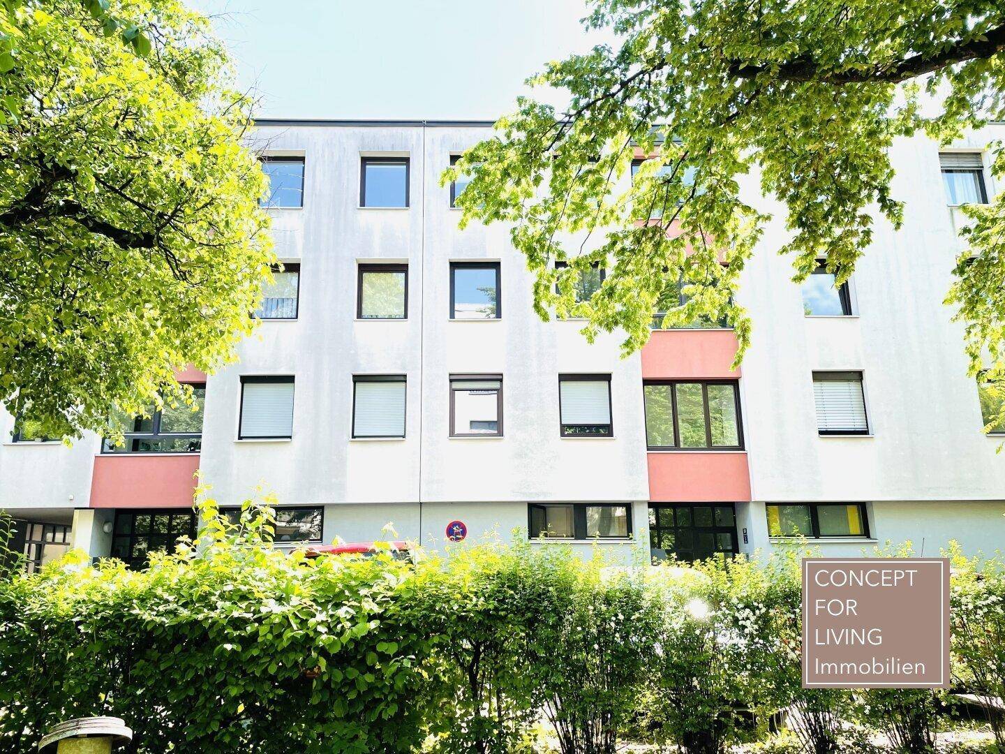 Mehrstöckiges Wohngebäude mit grüner Hecke und Bäumen im Vordergrund.