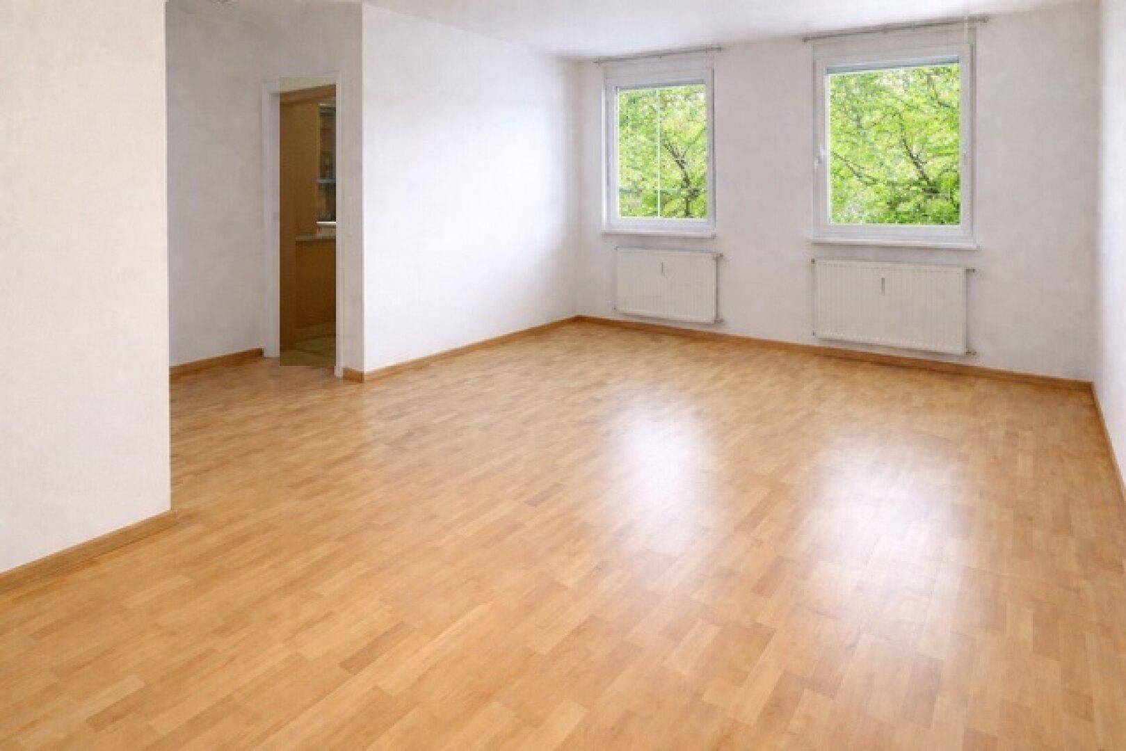 Geräumiges, leeres Zimmer mit Parkettboden und zwei Fenstern mit Blick ins Grüne.