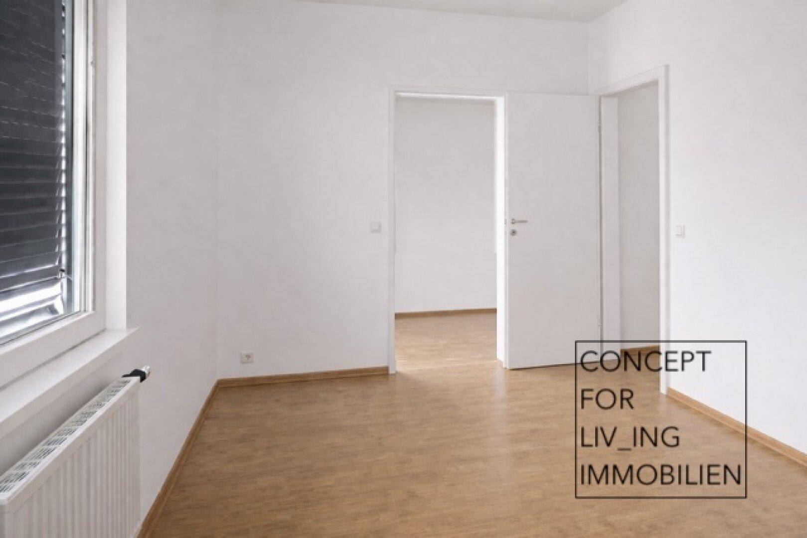 Helles, leeres Zimmer mit Parkettboden und Fenster, das zu weiteren Räumen führt.
