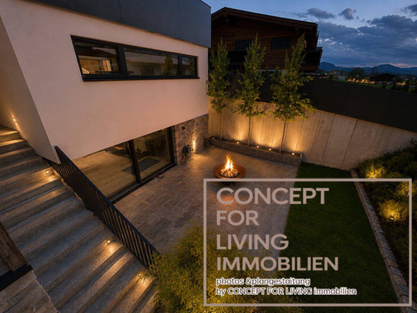 Beeindruckendes Anwesen bei Nacht mit beleuchteter Fassade, Garten und stimmungsvoller Feuerstelle.