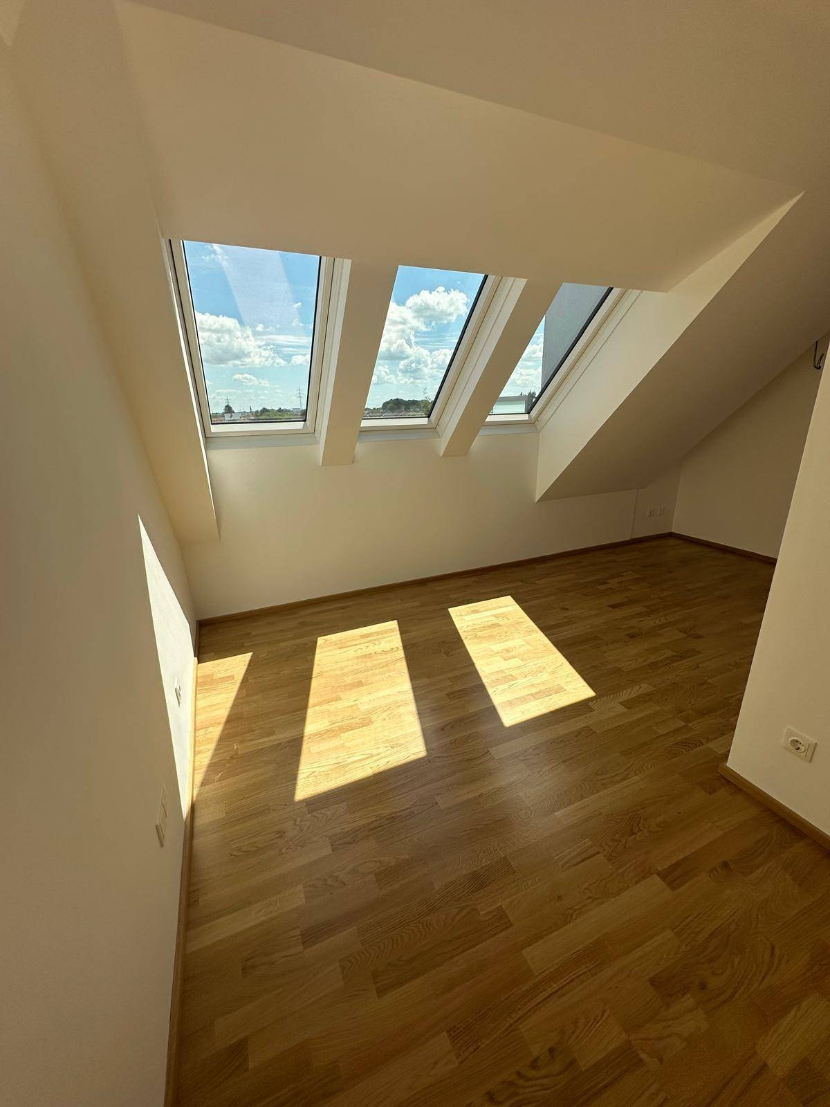 Ecke eines hellen Raumes mit drei Dachfenstern und glänzendem Parkettboden.
