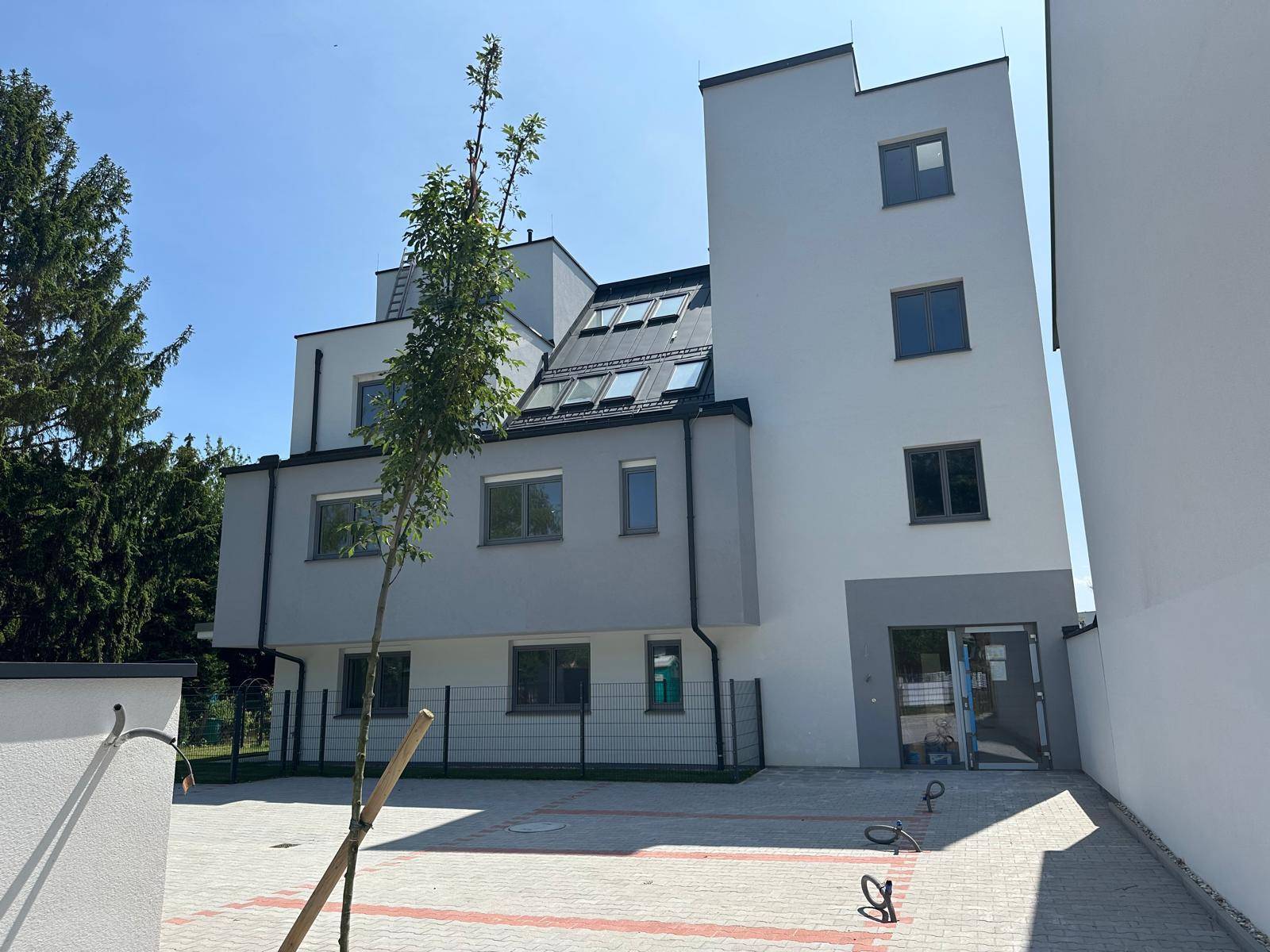 Modernes Neubaugebäude mit heller Fassade, großen Fenstern und einem gepflasterten Parkplatzbereich.