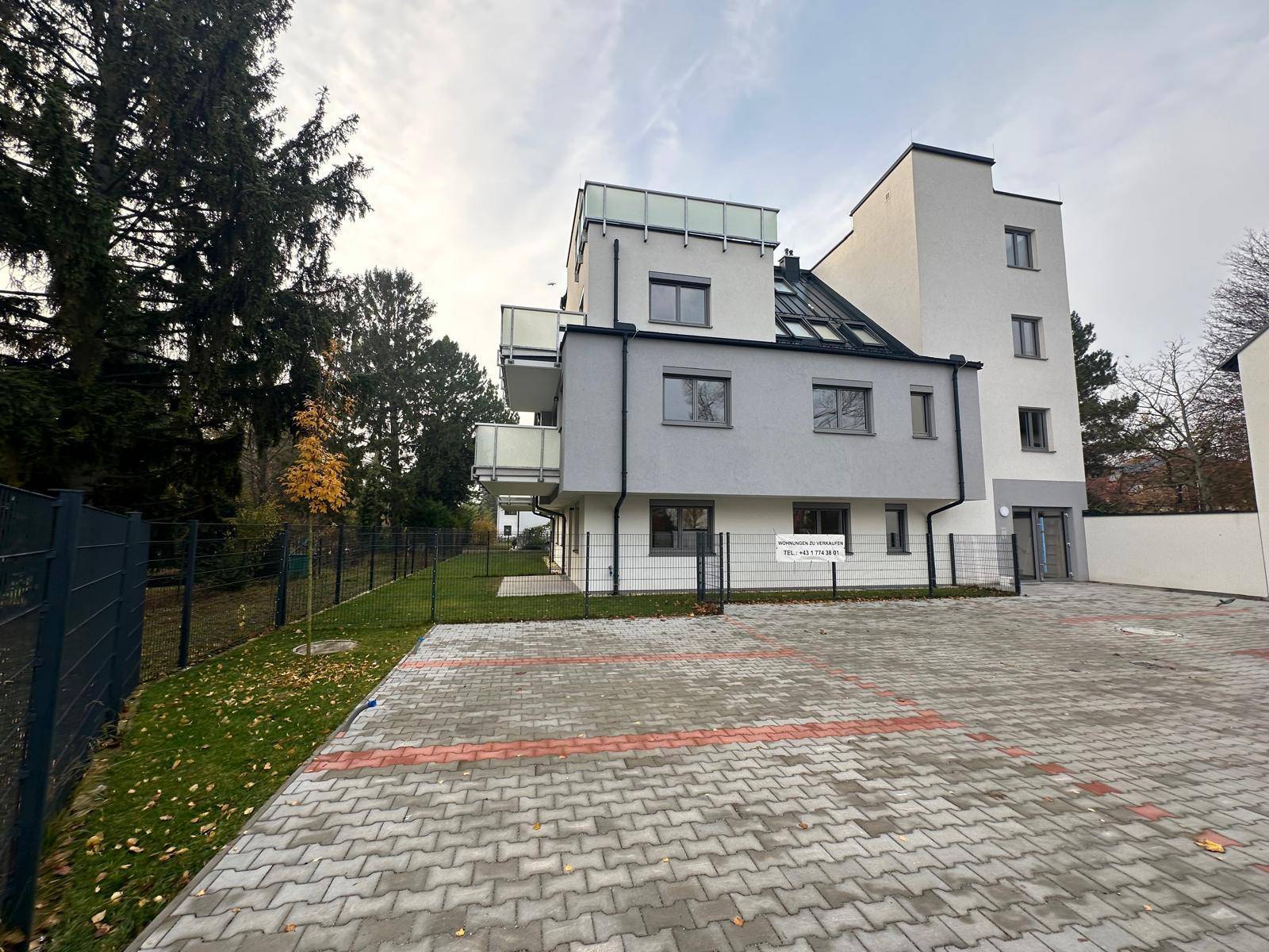 Neubau mit Parkplätzen und Zaun, umgeben von Bäumen unter bewölktem Himmel.