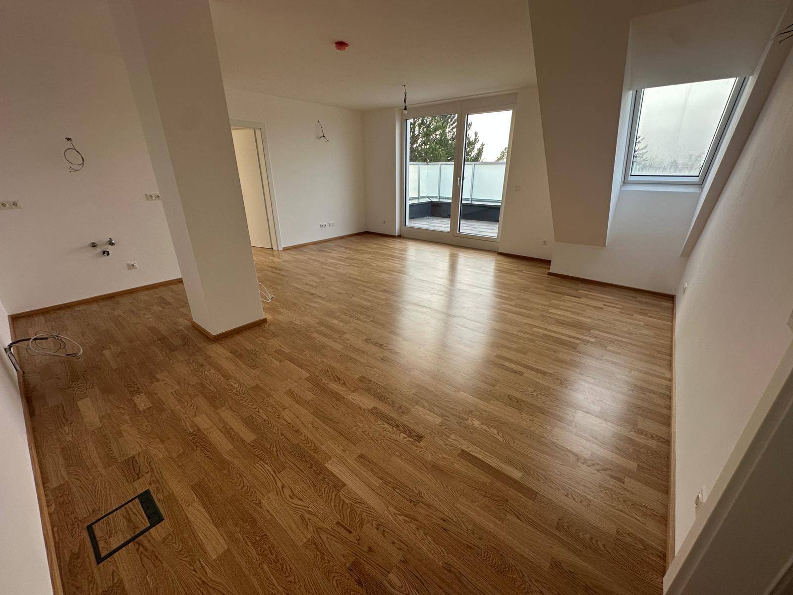Heller, leerer Wohnbereich mit Holzboden, Dachschräge und Zugang zu einem Balkon.