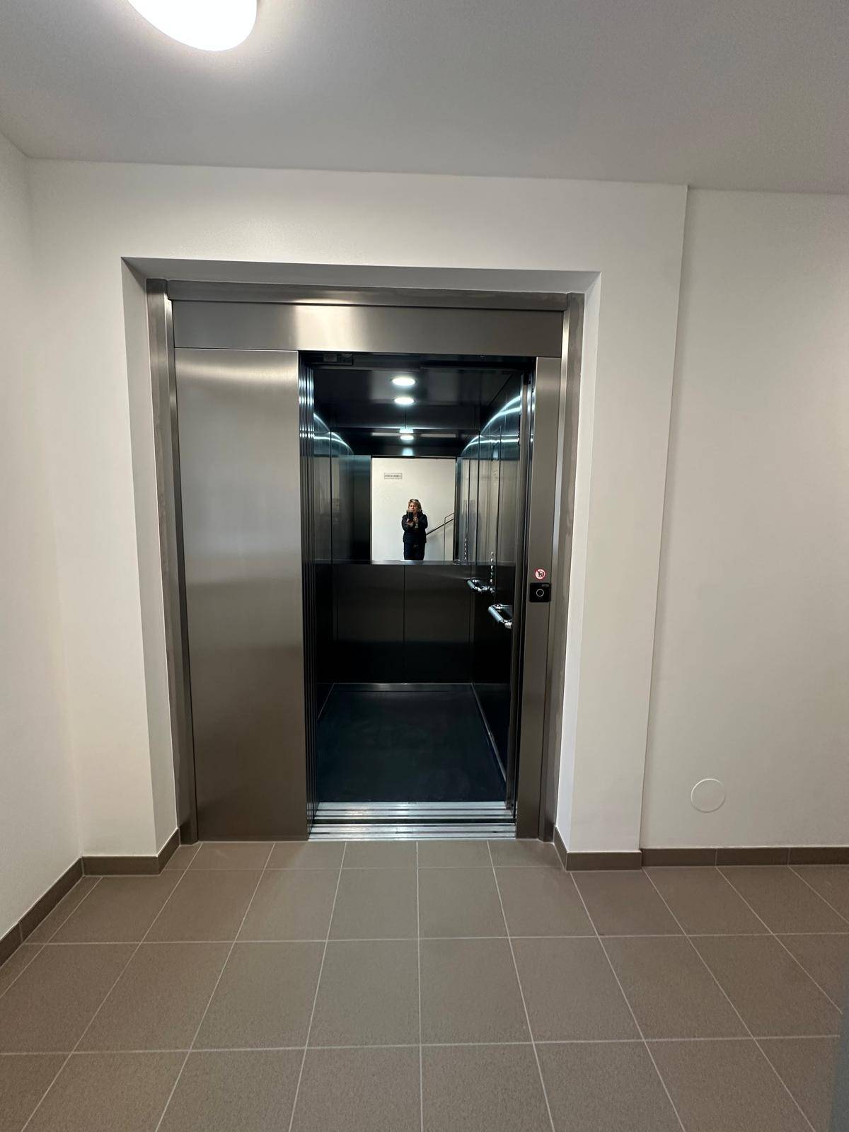 Moderner Aufzug in einem hellen Treppenhaus mit Fliesenboden.