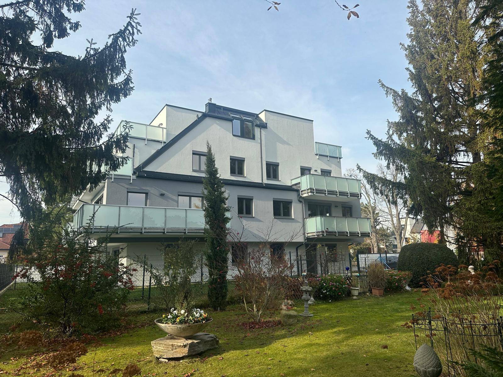 Gepflegter Gartenbereich vor der modernen Wohnanlage mit Balkonen und hohen Bäumen.