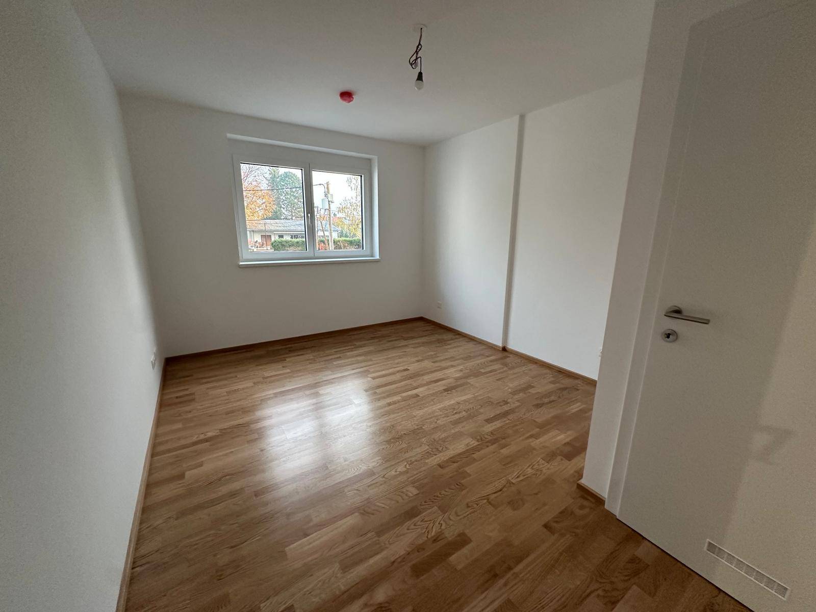 Ruhiges Schlafzimmer mit hellem Parkettboden und einem Fenster mit Blick ins Grüne.