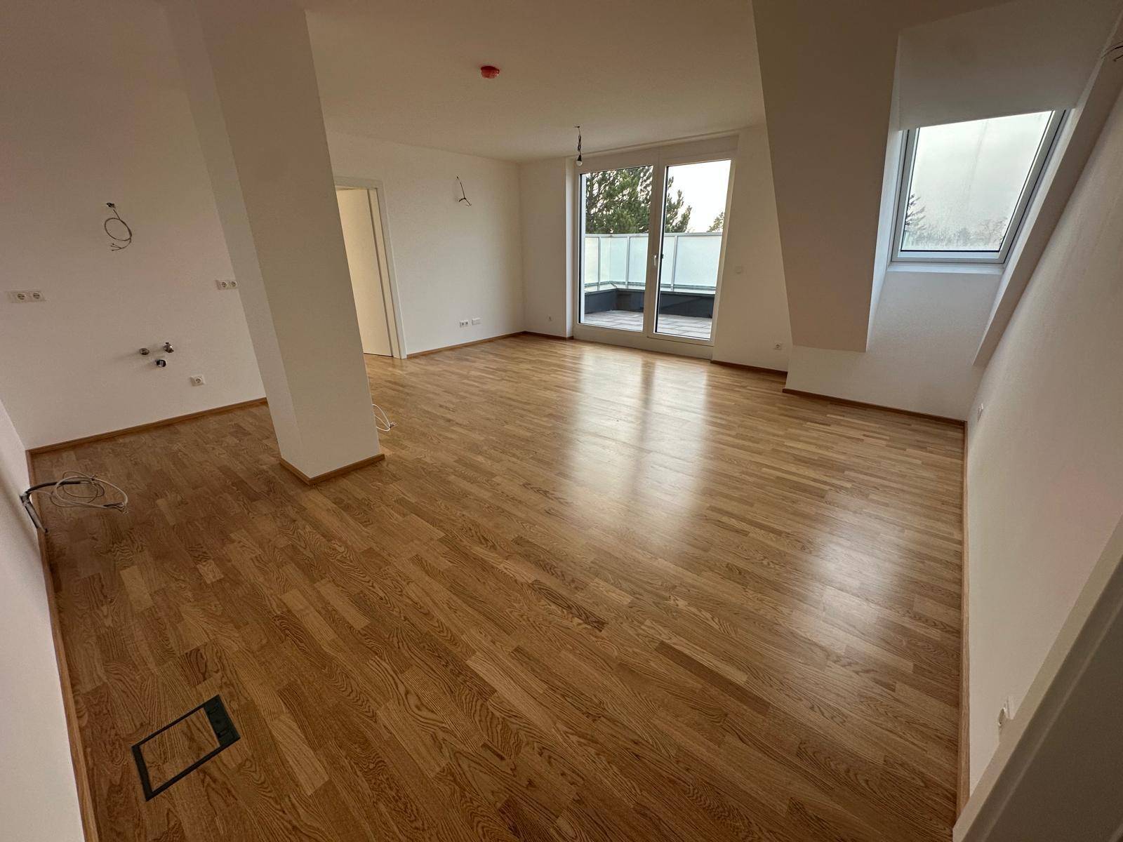 Heller, leerer Wohnbereich mit Holzboden, Dachschräge und Zugang zu einem Balkon.