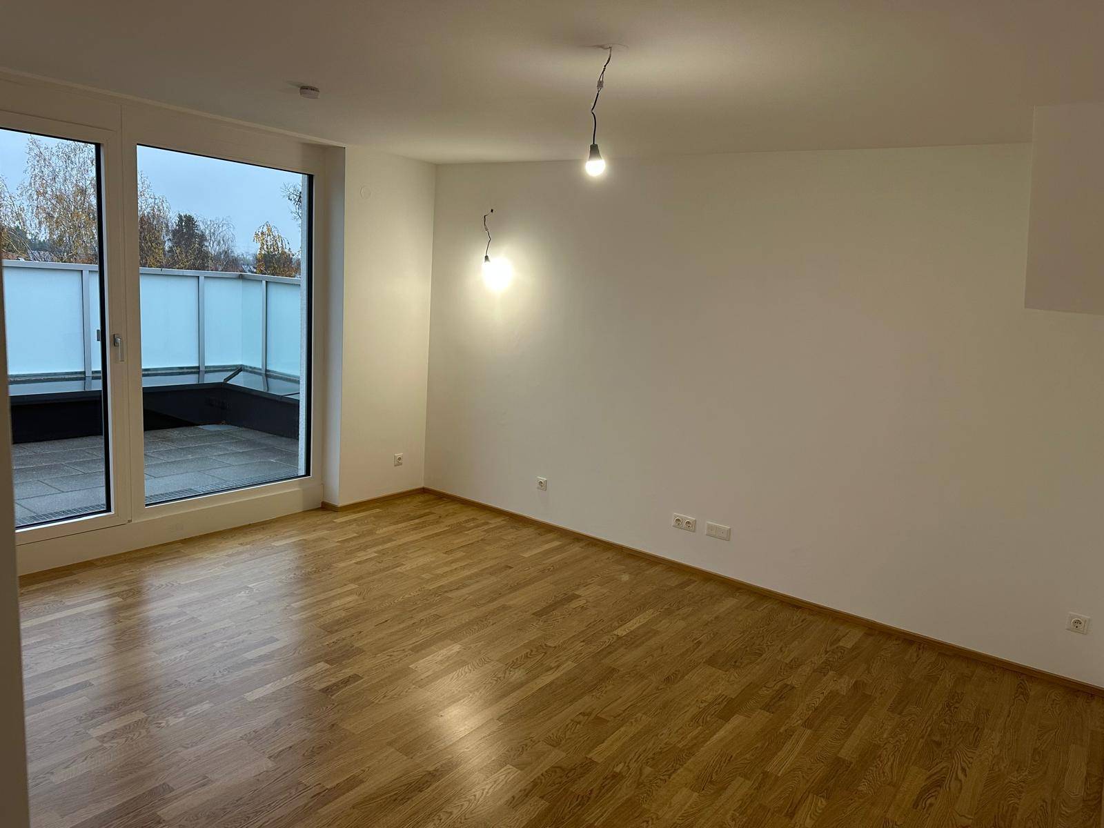 Helles Zimmer mit Holzboden und großer Schiebetür, die zu einer Außenterrasse führt.