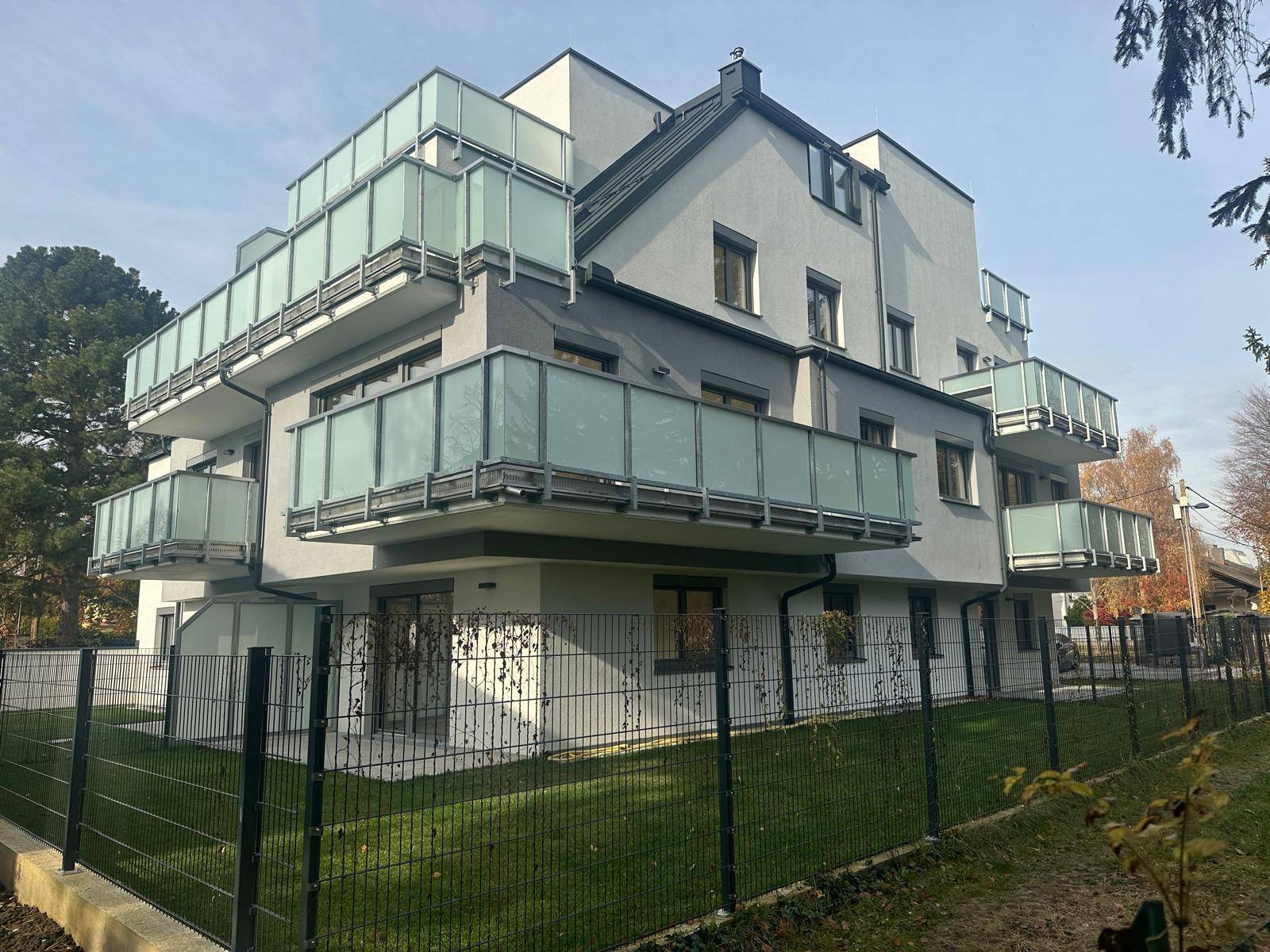 Modernes Mehrfamilienhaus mit vielen Balkonen, umzäuntem Rasen und klarem Himmel.