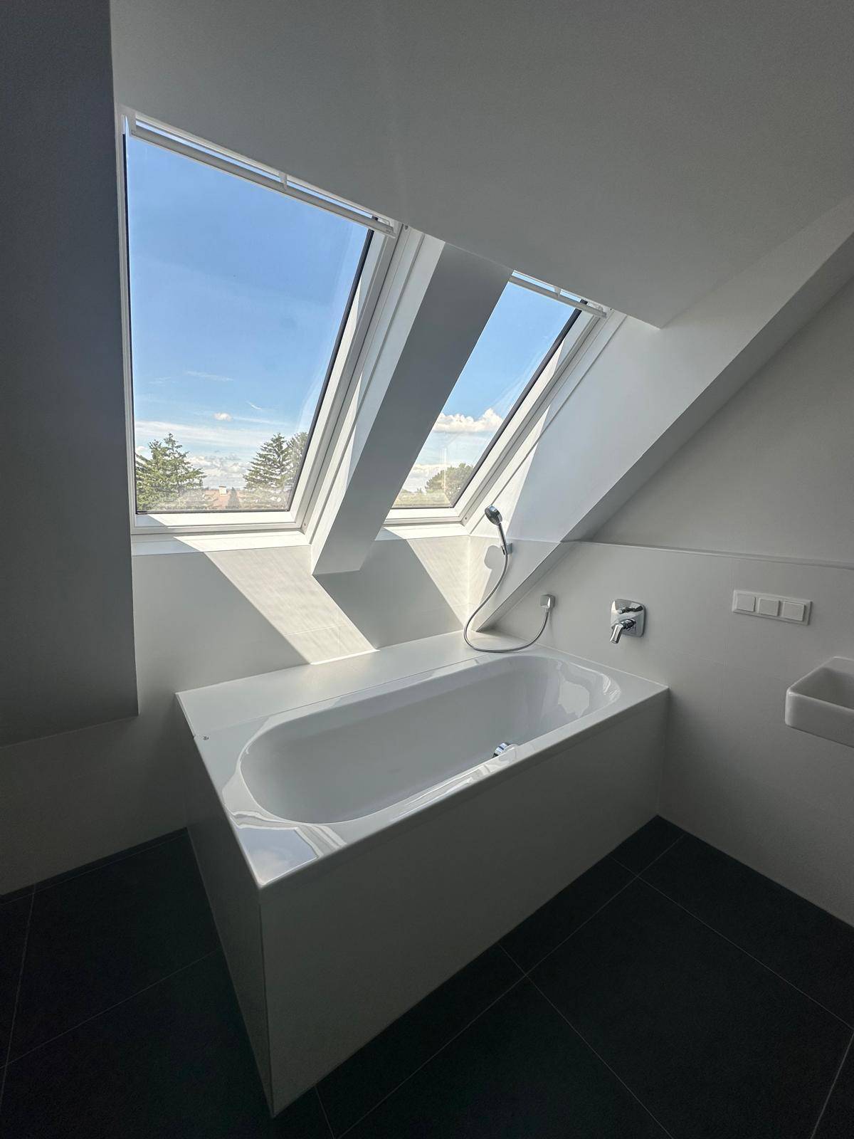 Helles Badezimmer mit Badewanne, Dachfenstern und dunklem Fliesenboden.