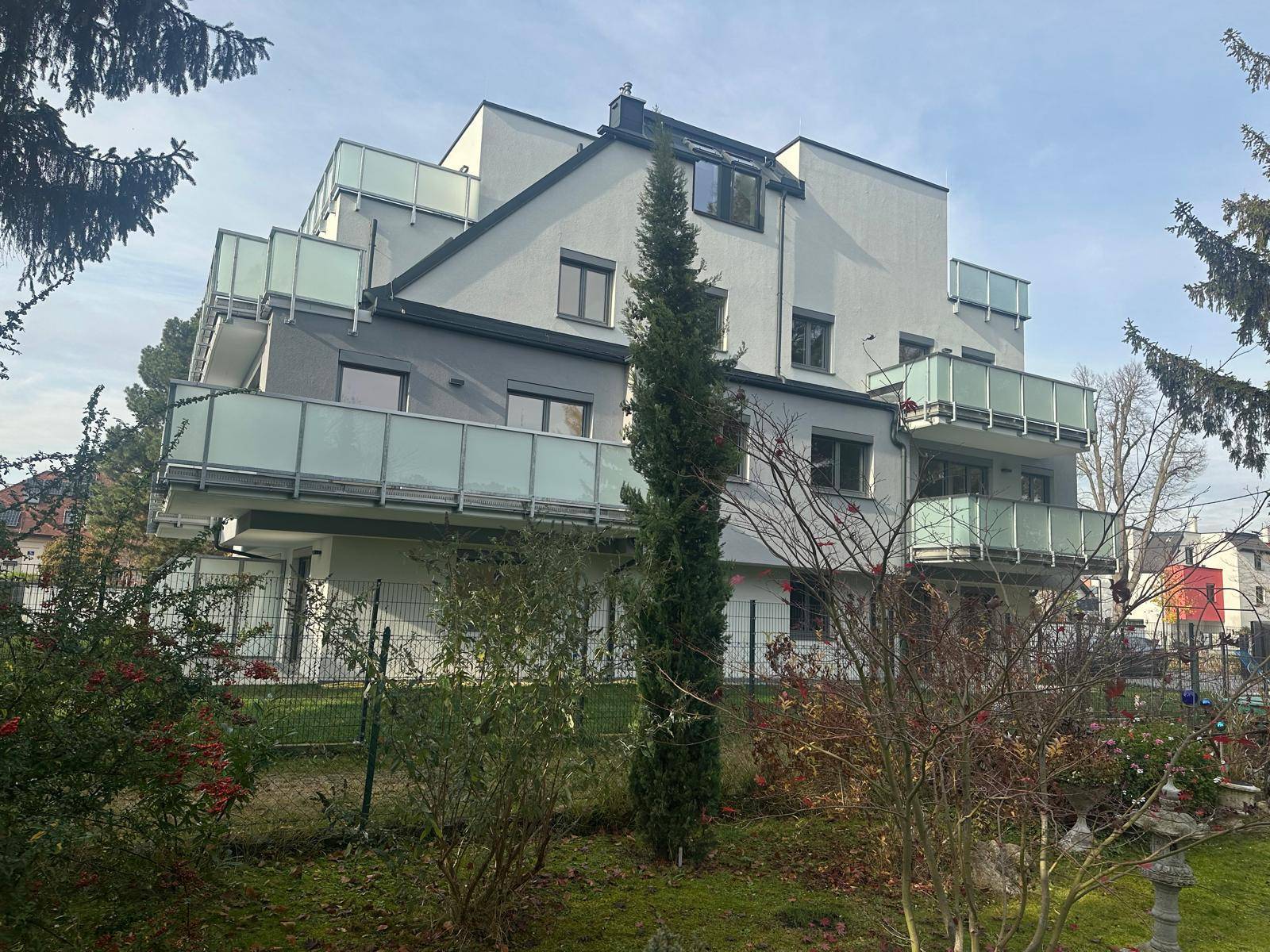 Ansicht eines modernen Wohngebäudes mit vielen Balkonen und einem grünen Garten mit Bäumen.