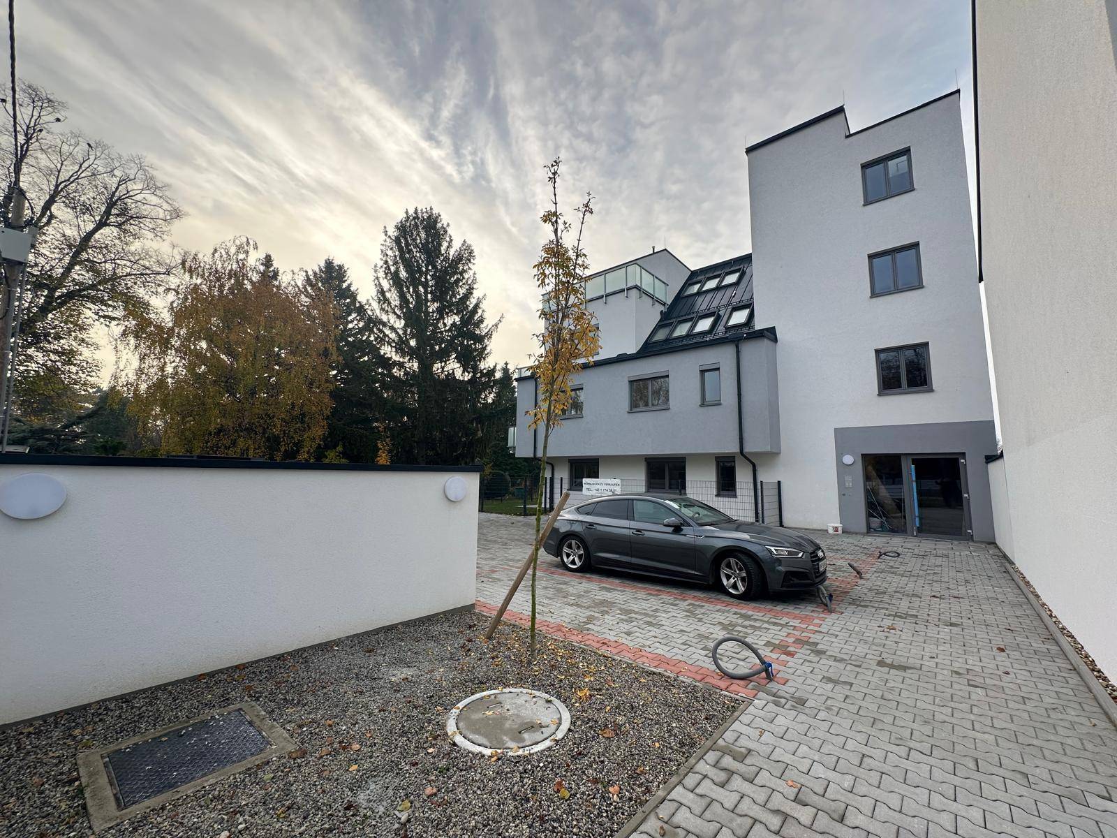 Modernes Mehrfamilienhaus mit Parkplatz und gepflasterter Zufahrt unter bewölktem Himmel.