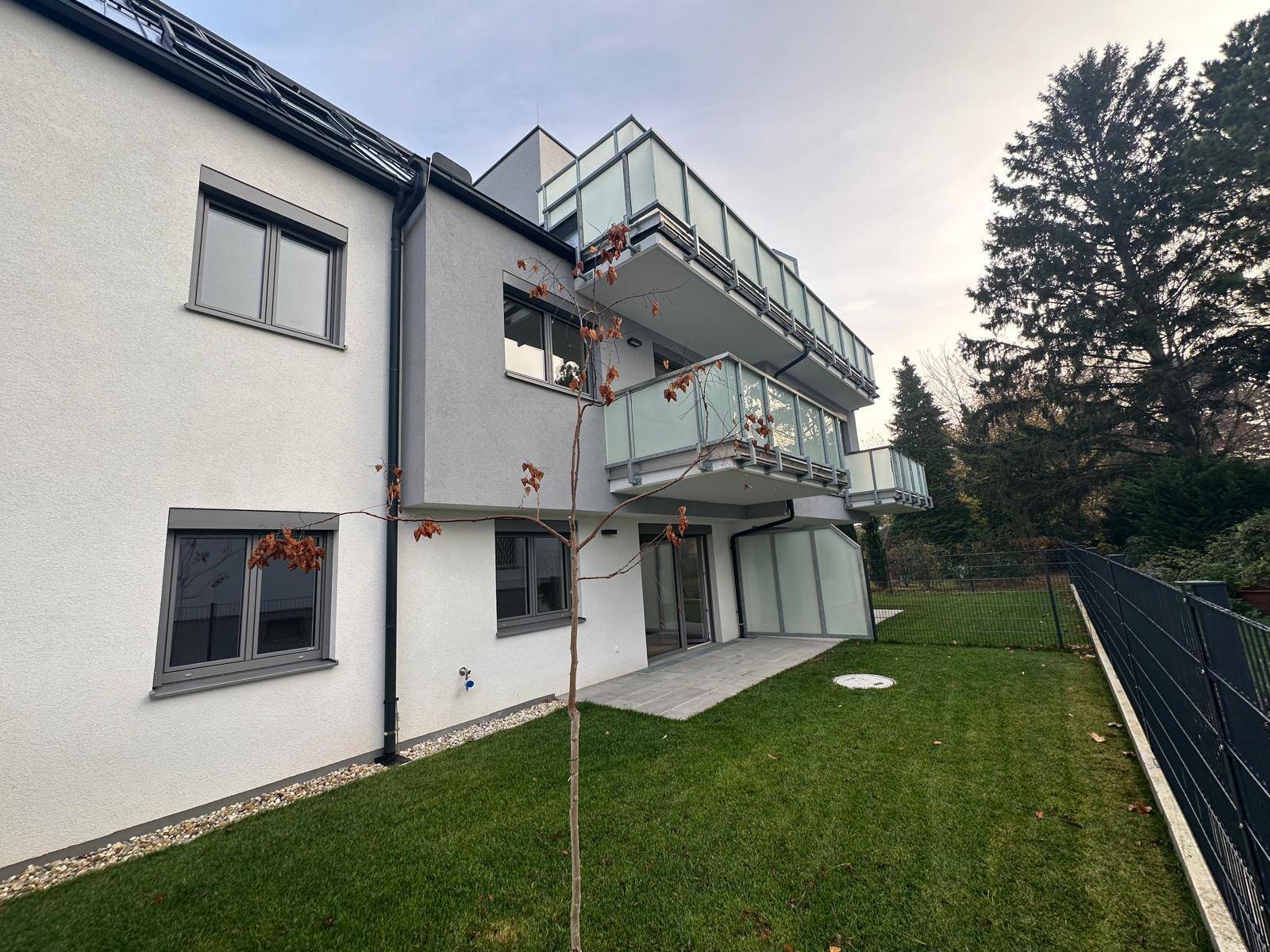 Seitenansicht des Neubaus mit Rasenfläche, Terrasse und Balkonen unter bewölktem Himmel.