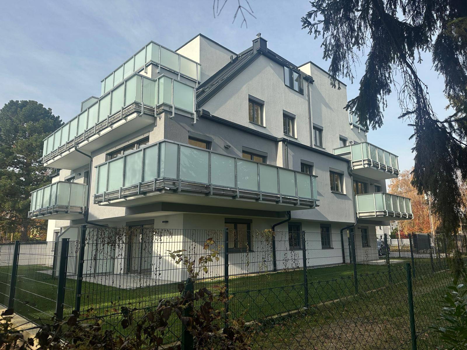 Modernes Wohngebäude von der Seite mit Balkonen, Zaun und Bäumen.