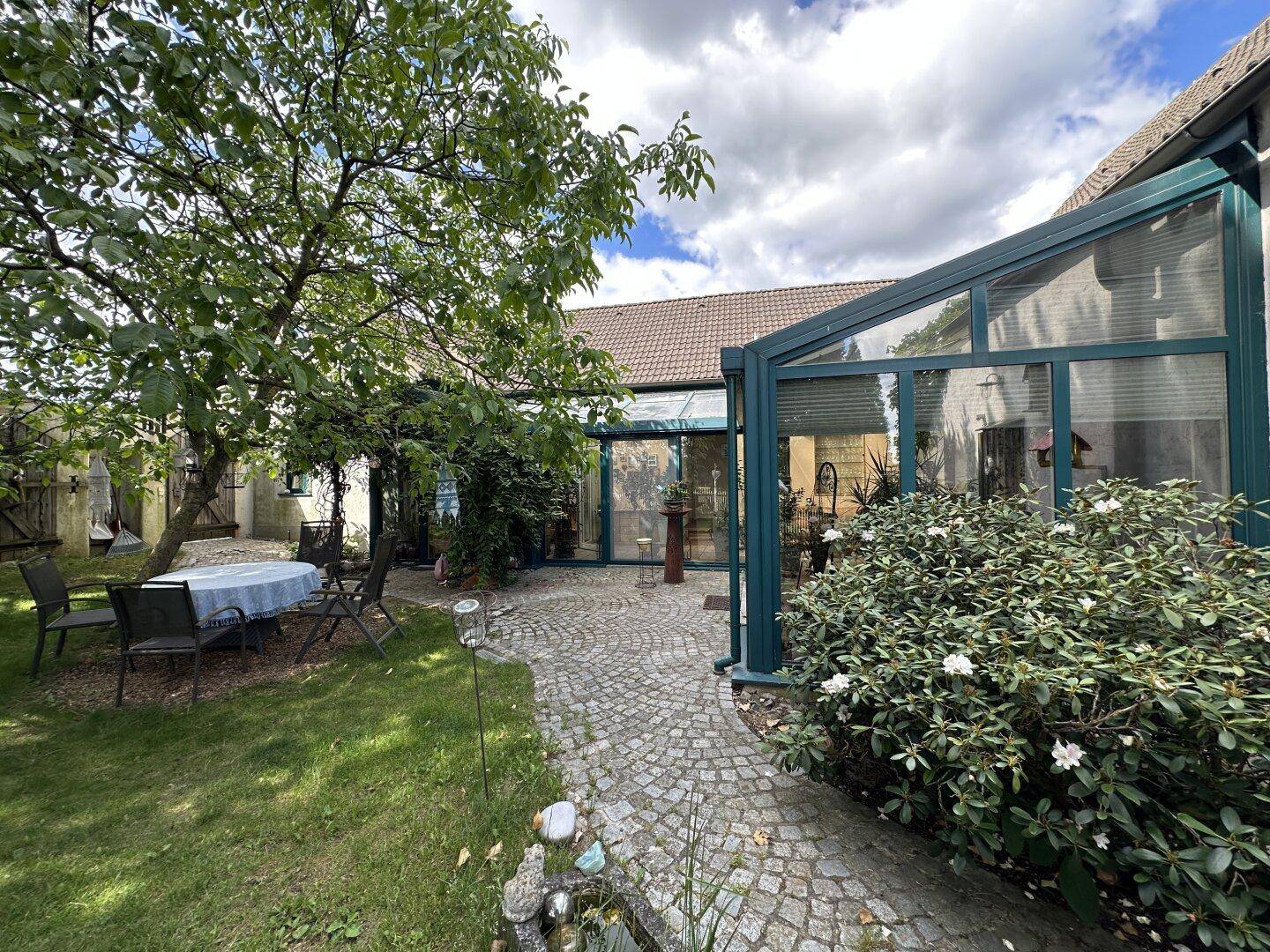 Idyllischer Gartenbereich mit Sitzgelegenheiten und einem Wintergarten, umgeben von Grün.
