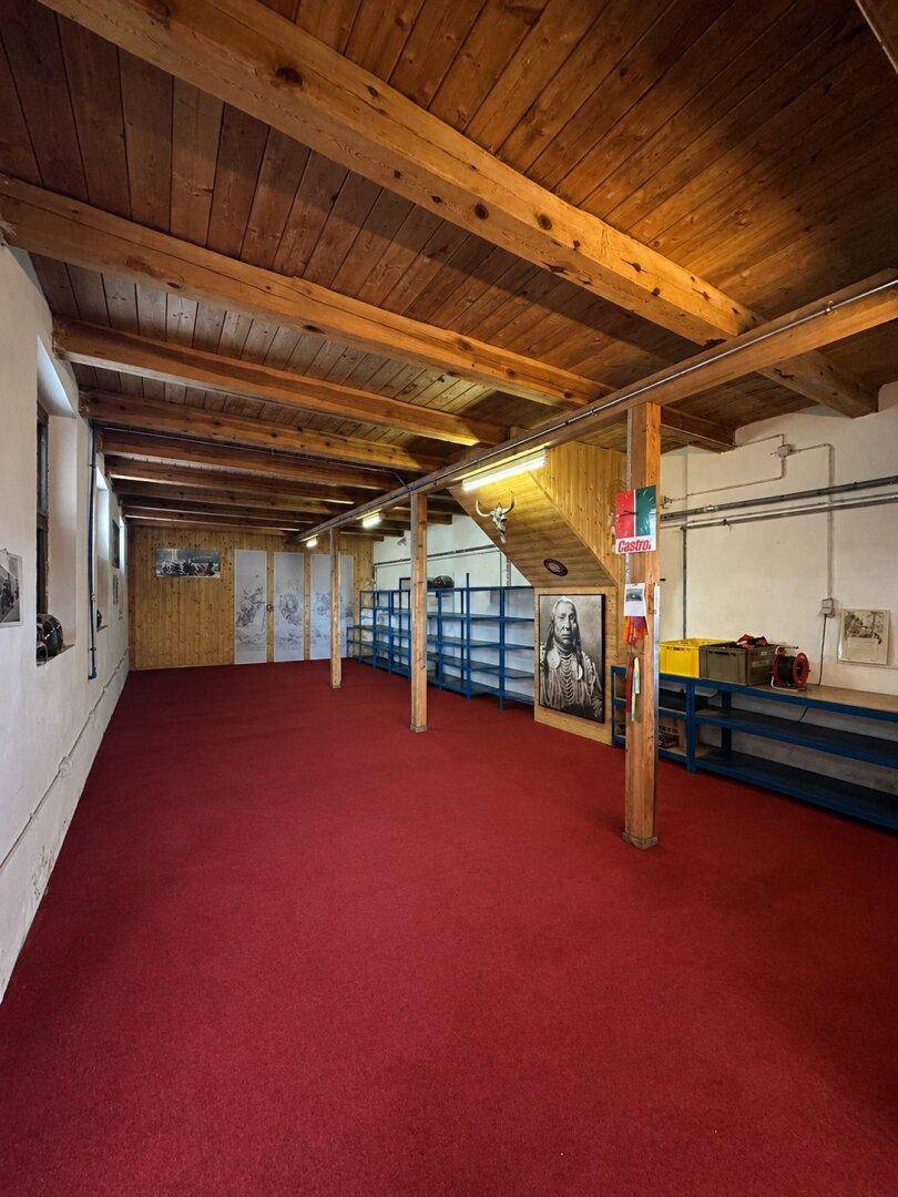 Großer Raum mit rotem Teppichboden, sichtbaren Holzbalken und Regalen an den Wänden.